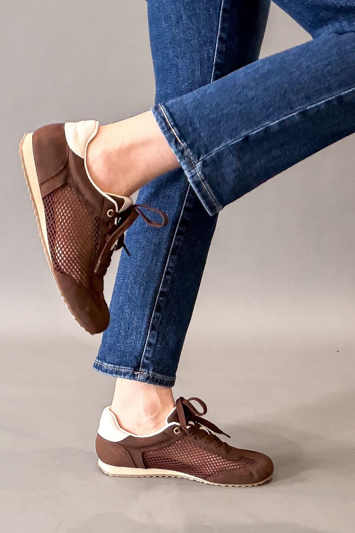 A person wearing brown mesh lace-up sneakers with blue jeans, shown from the knees down against a plain gray studio background.