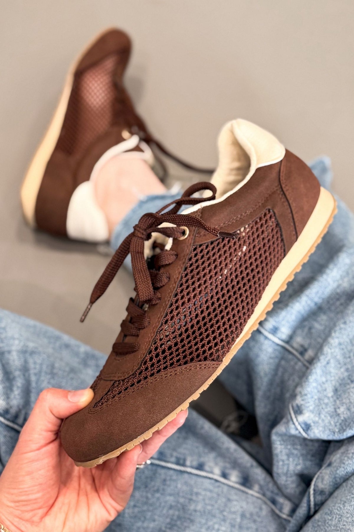 A person holds a brown mesh sneaker with a beige sole, with their jeans and the other shoe visible in the background.