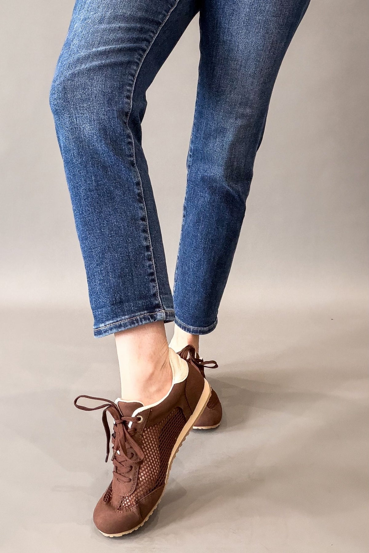 A person wearing blue jeans and brown lace-up sneakers, shown from the knees down against a plain light gray studio background.