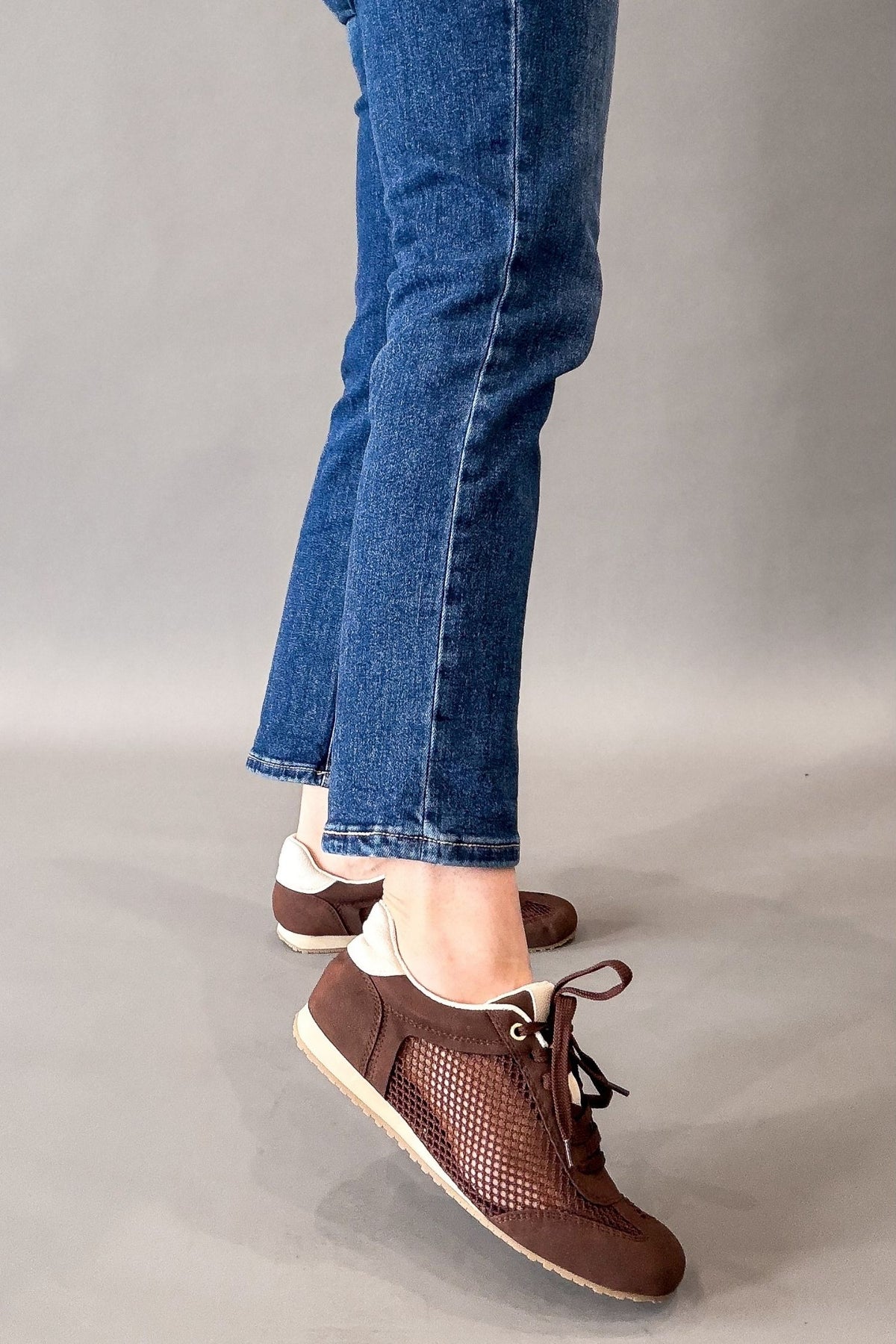 A person wearing brown lace-up sneakers with a textured mesh side panel and white soles, paired with blue jeans, shown from the knees down against a plain gray studio background.
