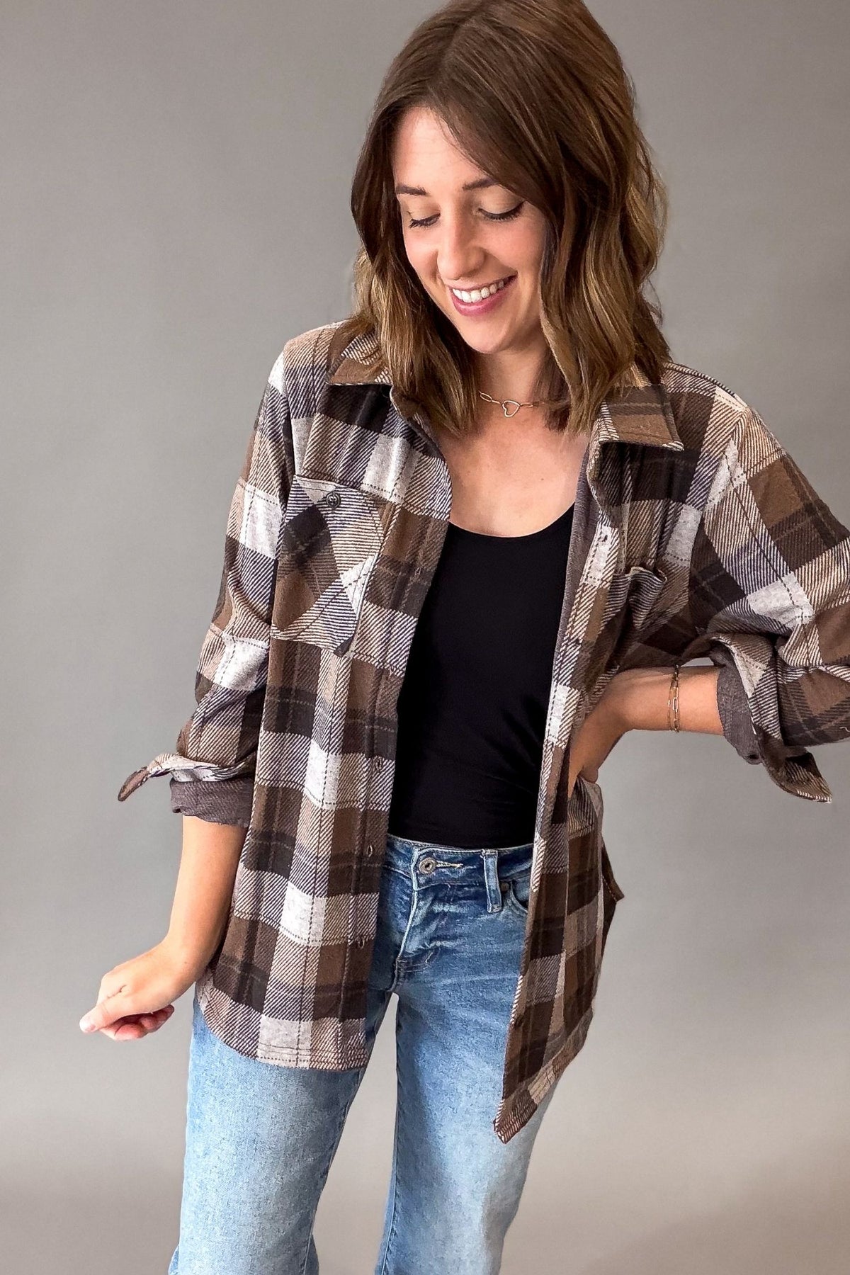 A woman wearing a brown, beige, and white plaid flannel shirt over a black tank top and blue jeans, standing against a plain gray studio background.