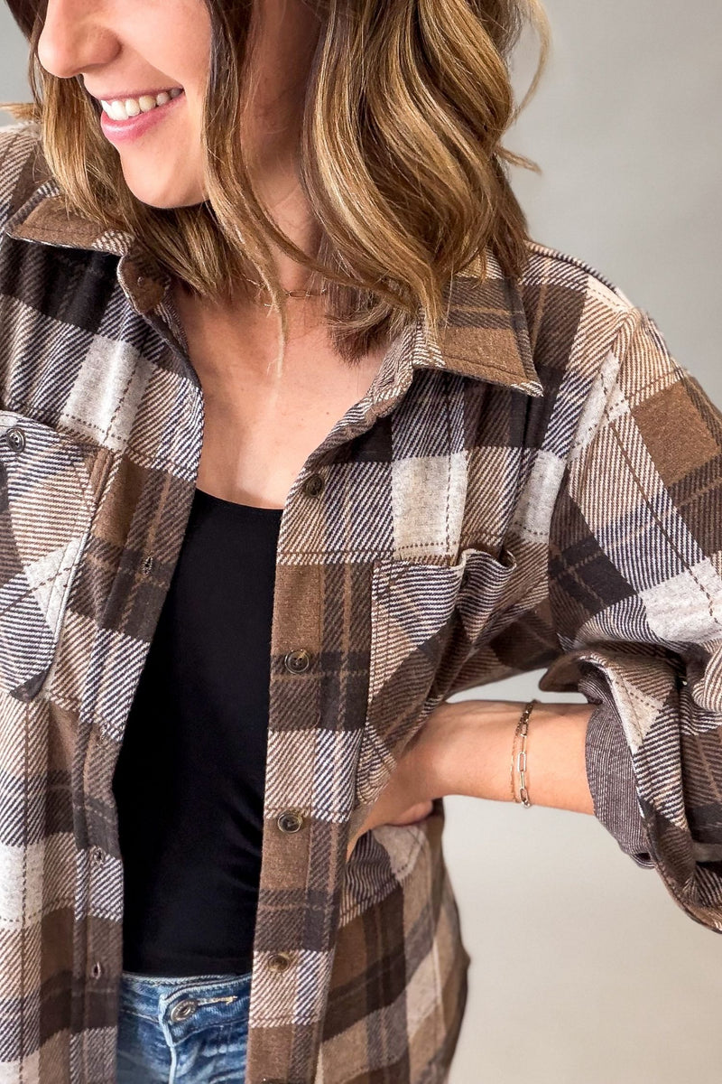 A woman wearing a brown, beige, and gray plaid button-up flannel shirt over a black tank top and blue jeans, shown in a close-up shot against a neutral gray background.