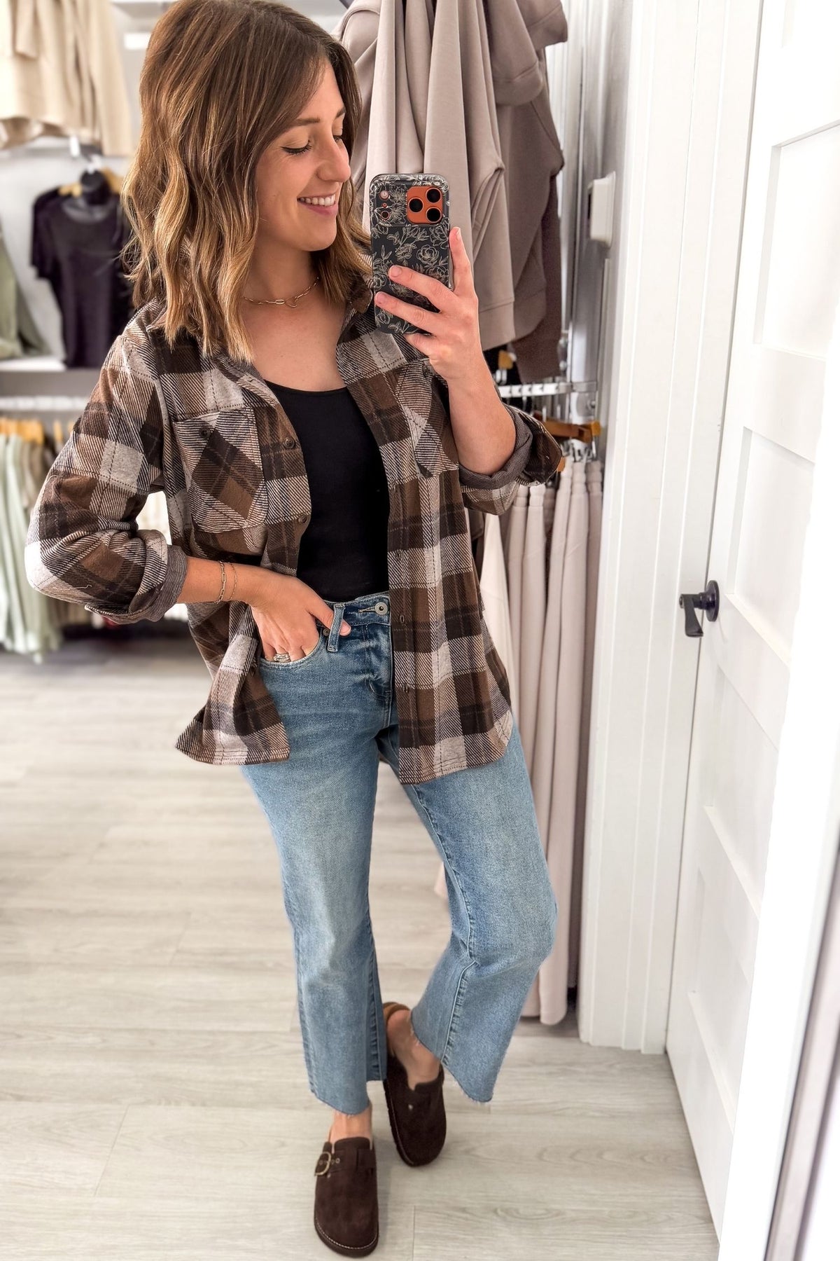 A woman takes a mirror selfie in a clothing boutique, wearing a brown and beige plaid flannel shirt over a black tank top, light-wash blue jeans, and brown slip-on loafers.