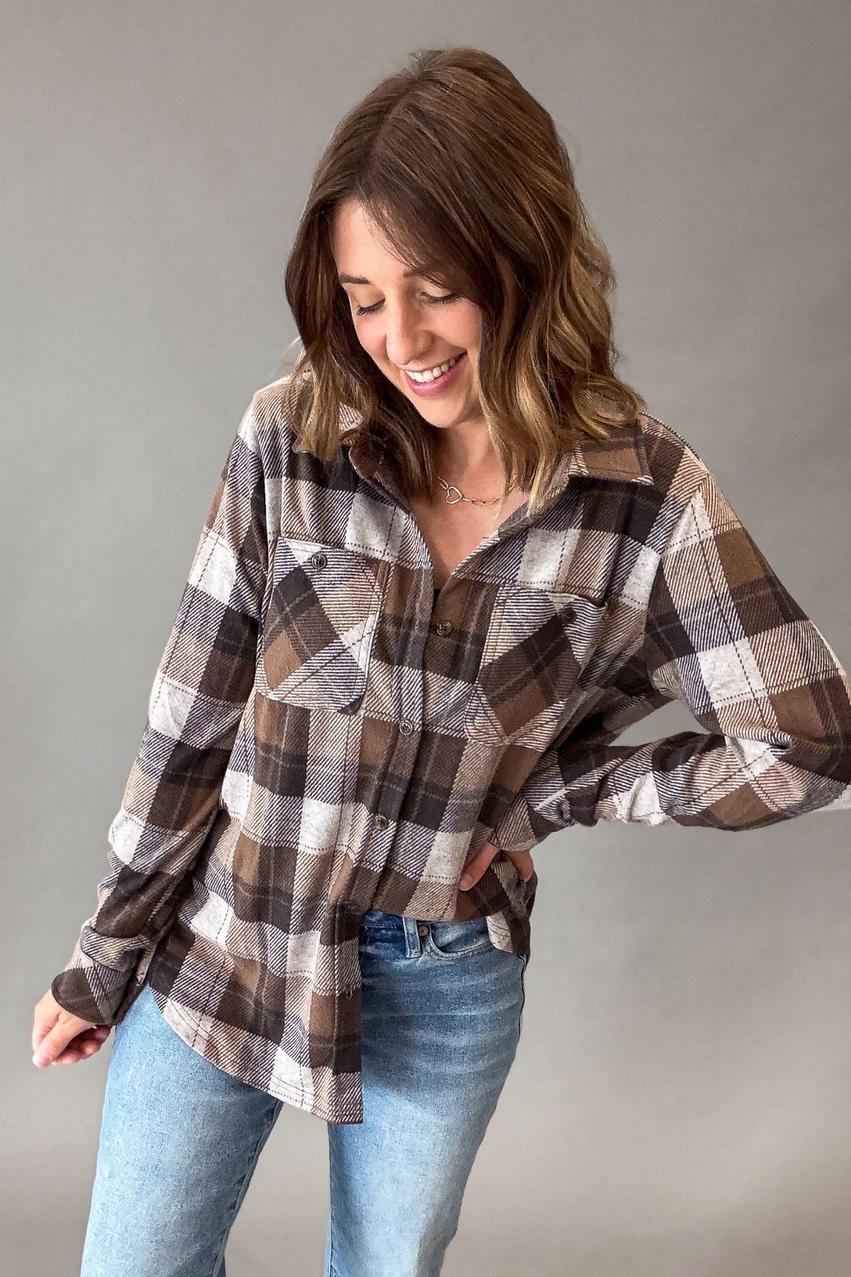 A woman wearing a brown, beige, and white plaid flannel button-up shirt with blue jeans, posing against a neutral gray studio background.