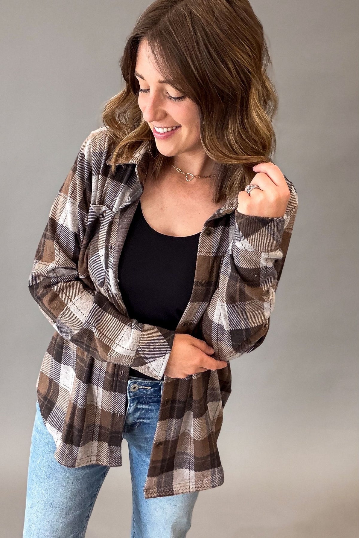 A woman wearing a brown, beige, and gray plaid button-up flannel shirt over a black tank top and blue jeans, posing against a neutral gray studio background.