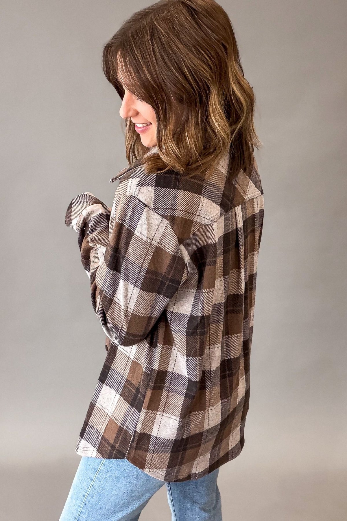 A woman wearing a brown, beige, and gray plaid flannel shirt, shown from a side-back angle against a neutral gray studio background.