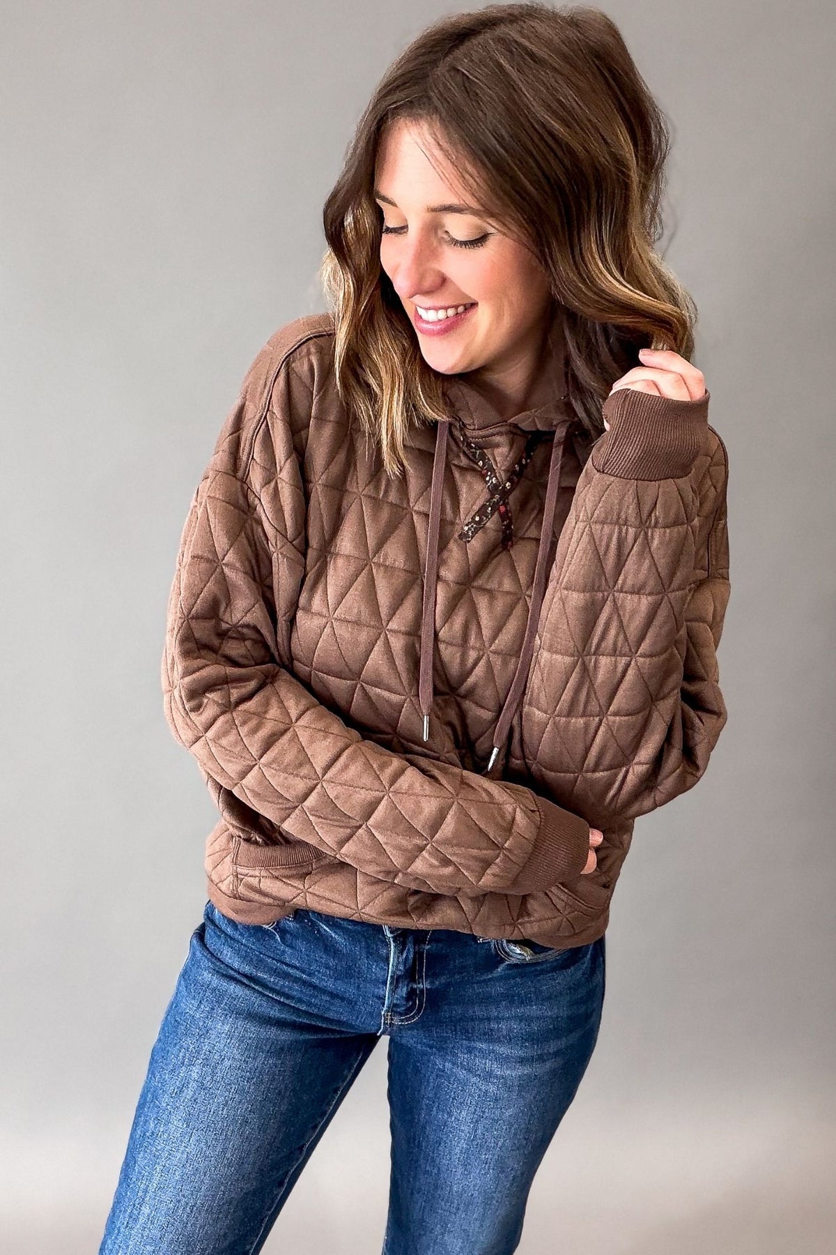 A woman wearing a brown diamond-quilted pullover hoodie with drawstrings and ribbed cuffs, paired with blue jeans, posing against a neutral gray studio background.