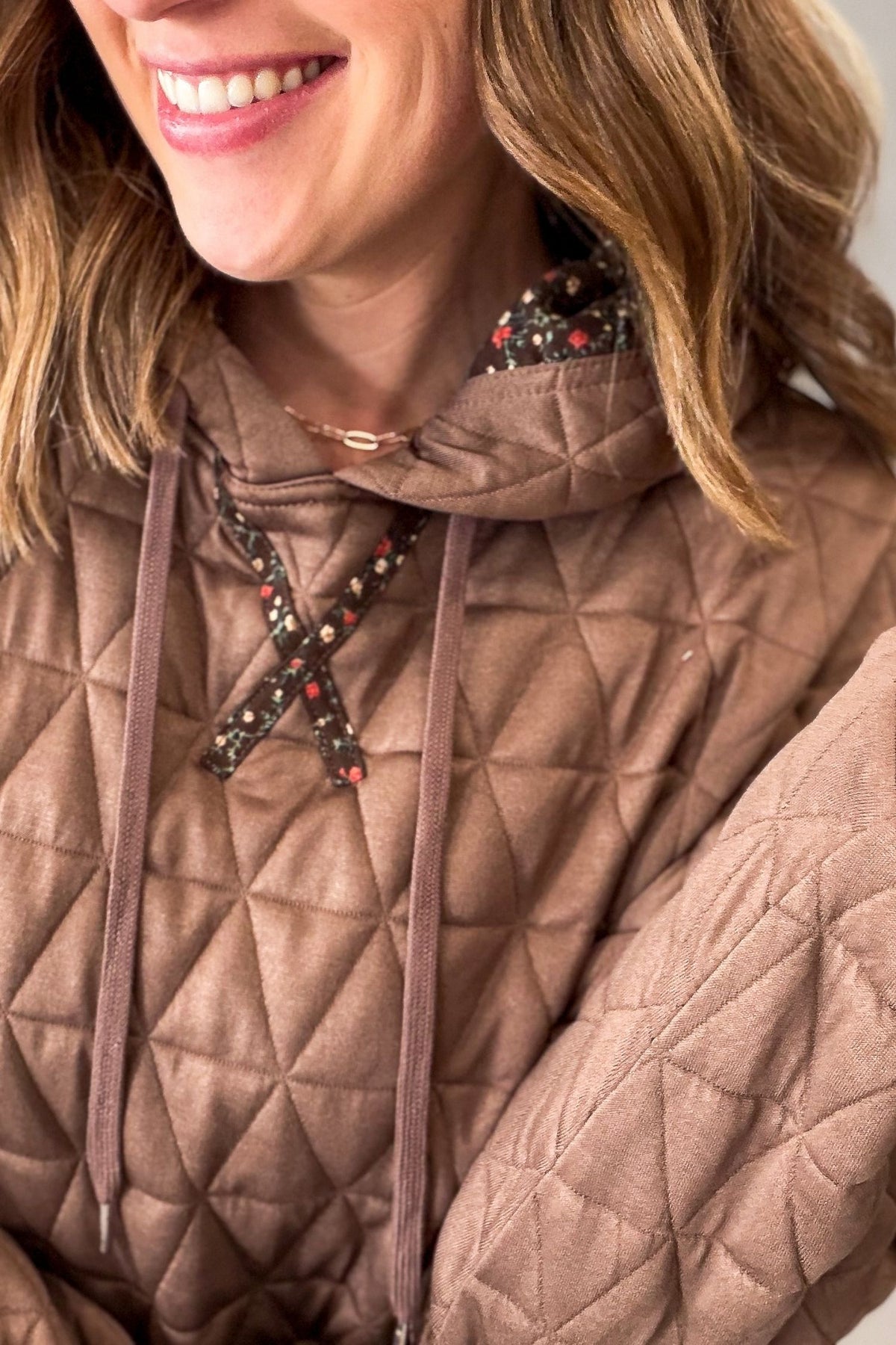 Close-up of a woman wearing a brown quilted hoodie with a geometric diamond pattern, matching drawstrings, and a decorative floral lace-up detail on the chest.