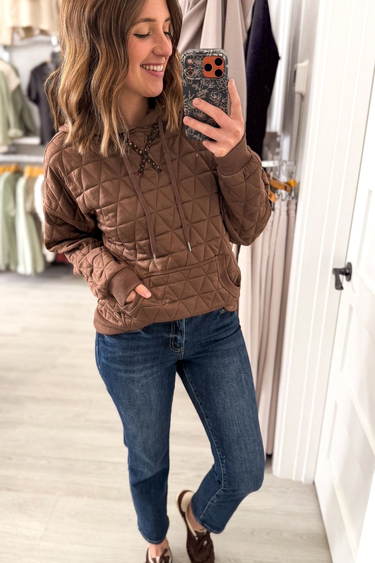 A woman takes a mirror selfie in a boutique wearing a brown diamond-quilted hoodie with lace-up detailing at the neck, paired with blue jeans and brown loafers.