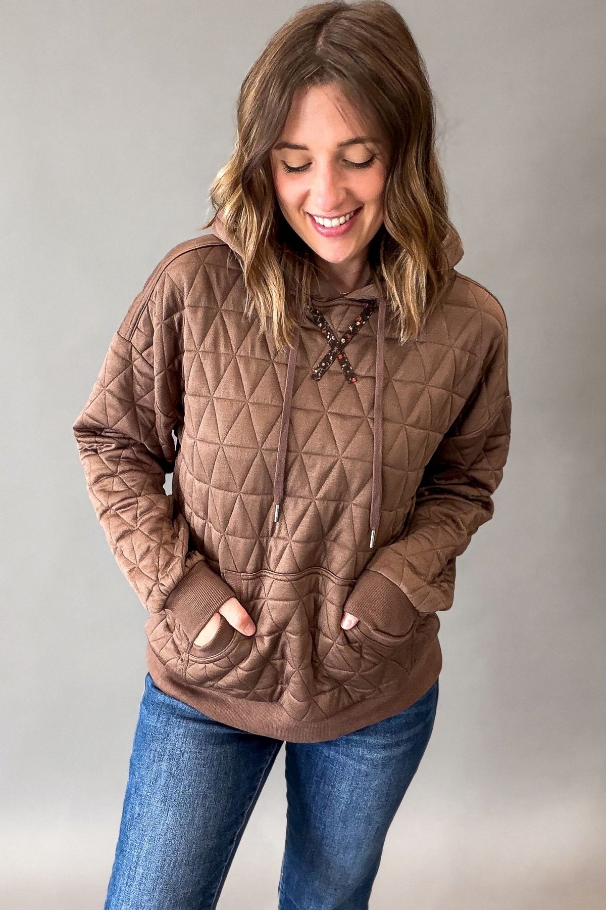 A woman wearing a brown quilted pullover hoodie with lace-up detail at the neckline and front pockets, paired with blue jeans, standing against a neutral gray studio background.