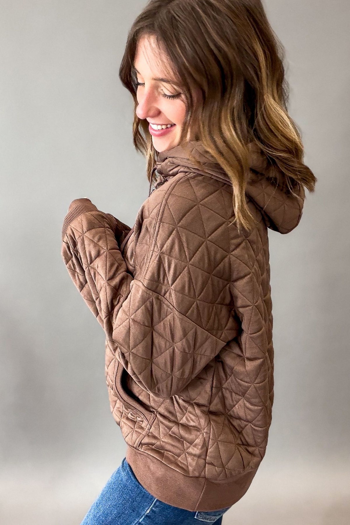 A woman wearing a brown quilted hooded jacket, shown from a side-back angle against a plain gray studio background.