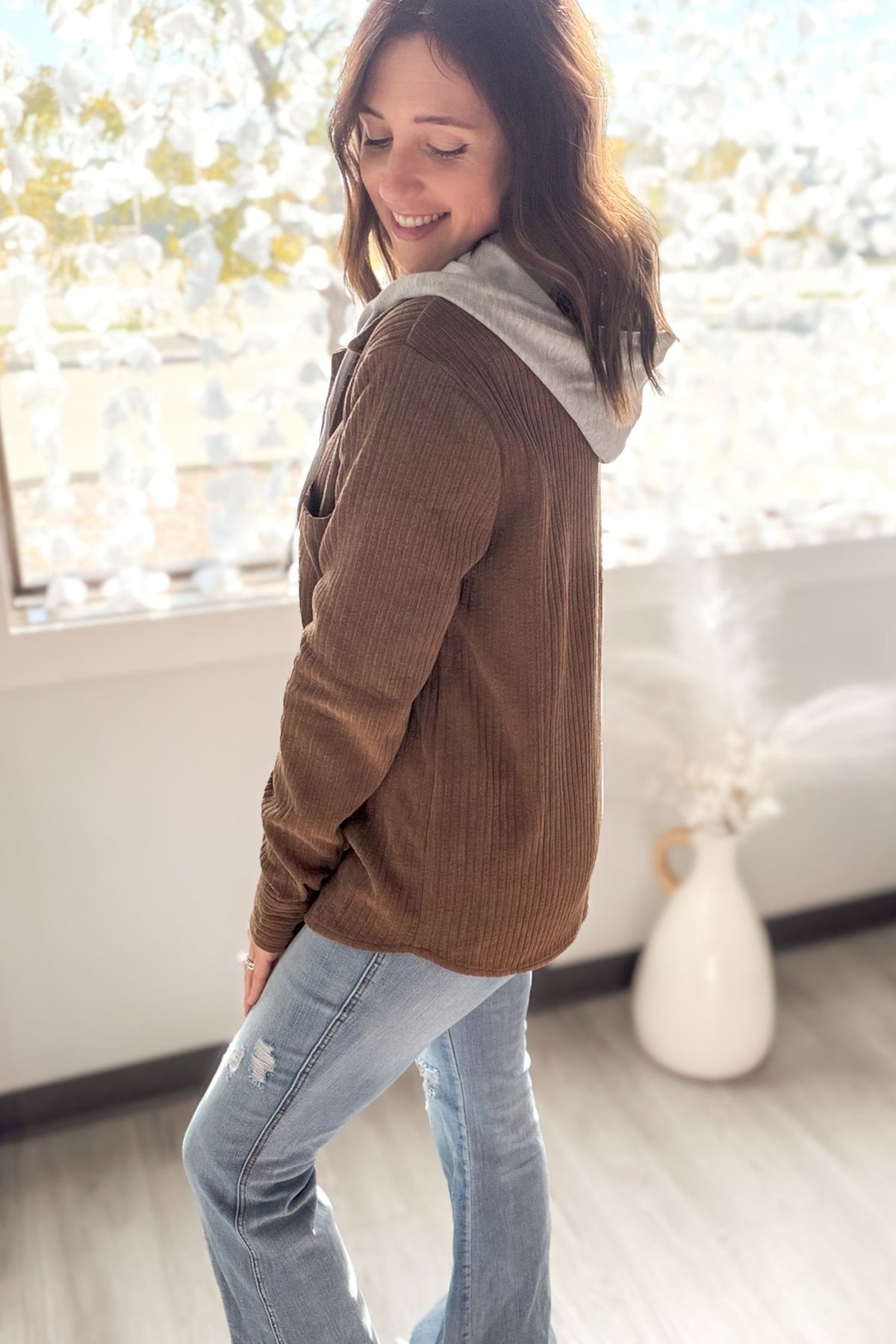 Woman wearing a brown/camel jacket with a hood in a room with large windows.