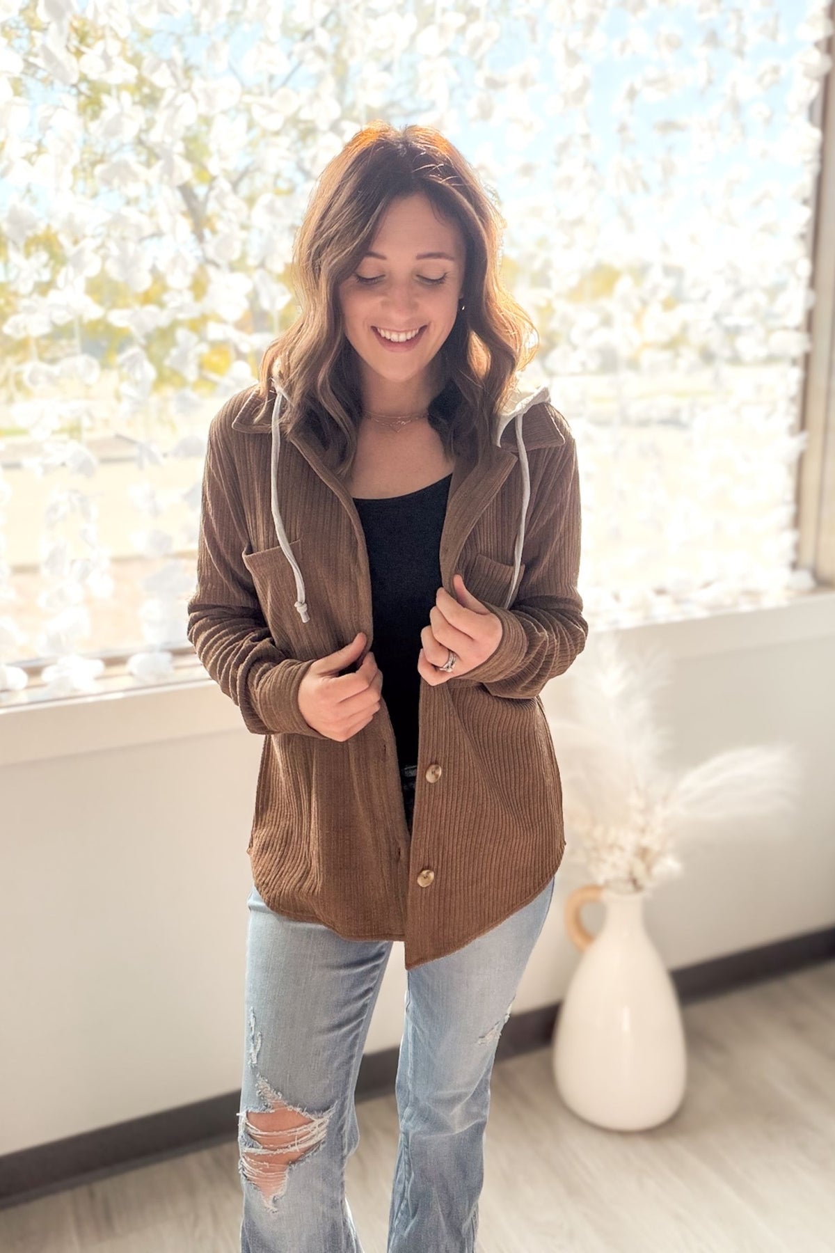 Woman wearing a brown/camel jacket and blue jeans standing in a room with a window in the background.