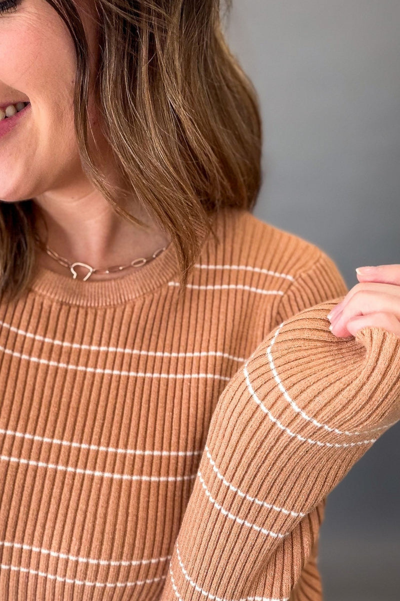 A close-up of a woman wearing a ribbed crewneck sweater in a warm camel brown with thin white horizontal stripes. She is smiling, and her hand is touching the sleeve of the sweater.