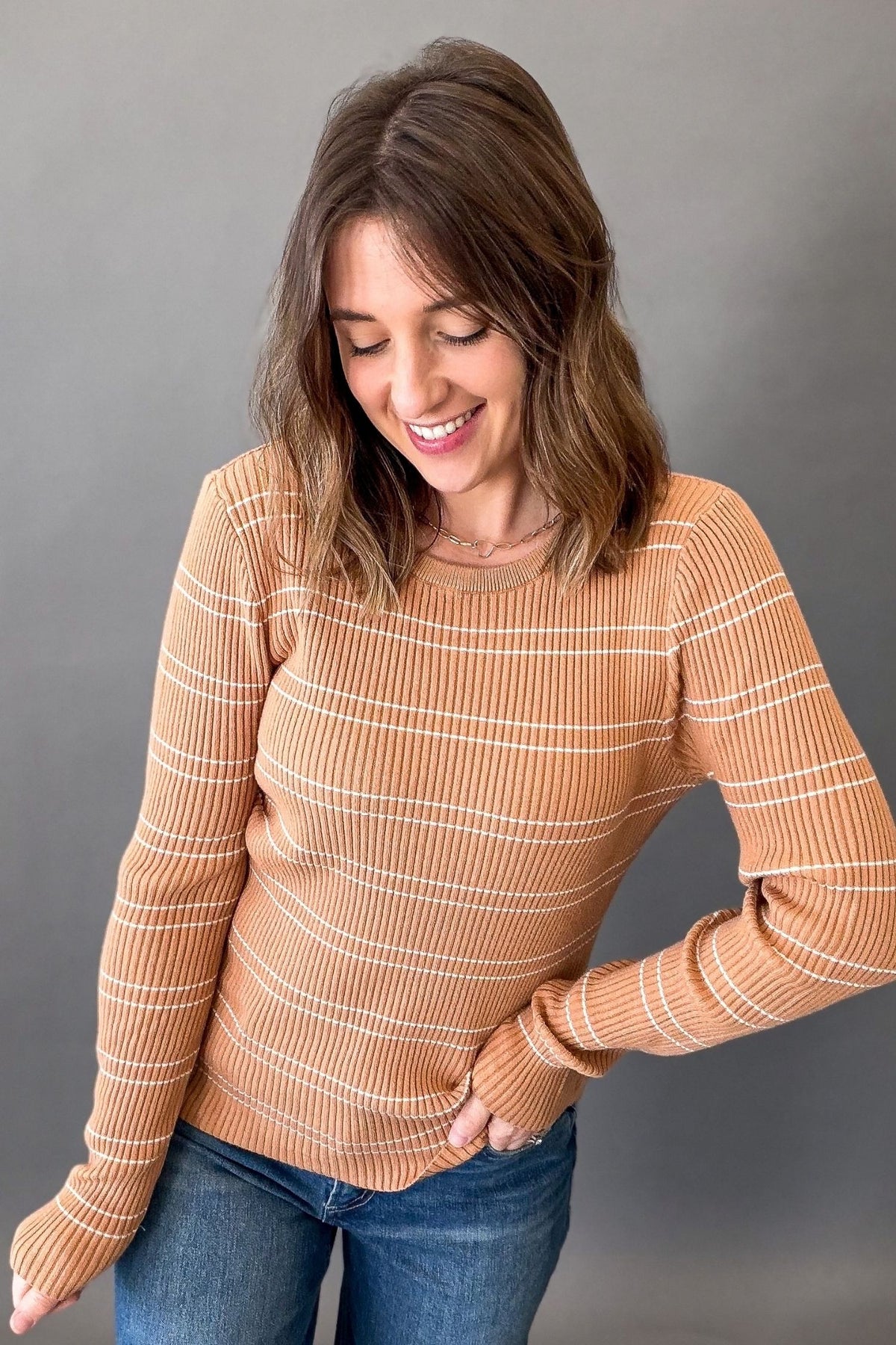 A woman wearing a ribbed long-sleeve crewneck sweater in a warm tan color with thin white horizontal stripes, paired with blue jeans, against a solid gray background.