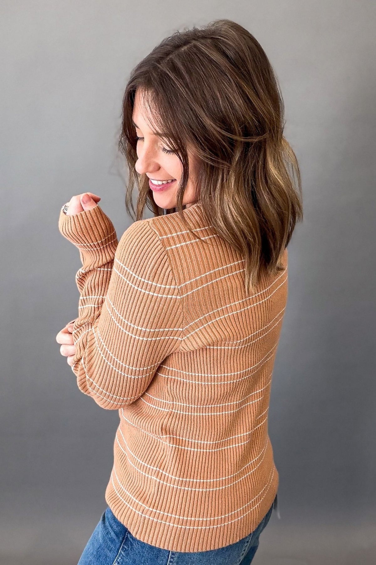 A woman wearing a ribbed knit turtleneck sweater in a warm tan color with thin white horizontal stripes, paired with blue jeans, photographed against a solid gray studio background.