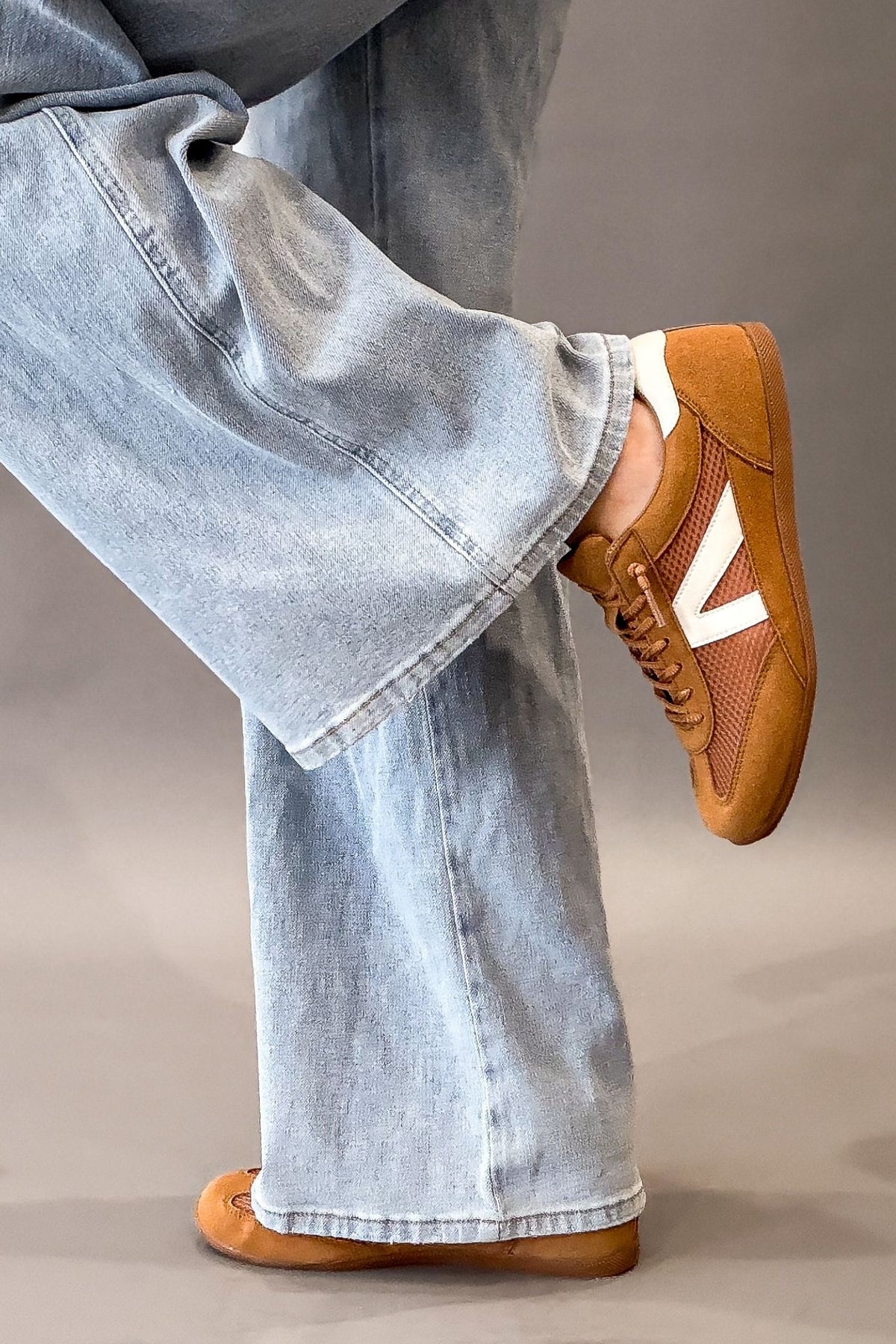 A person wearing light-wash wide-leg jeans and brown faux suede sneakers with a white V-shaped side stripe, shown from the waist down against a neutral studio background.