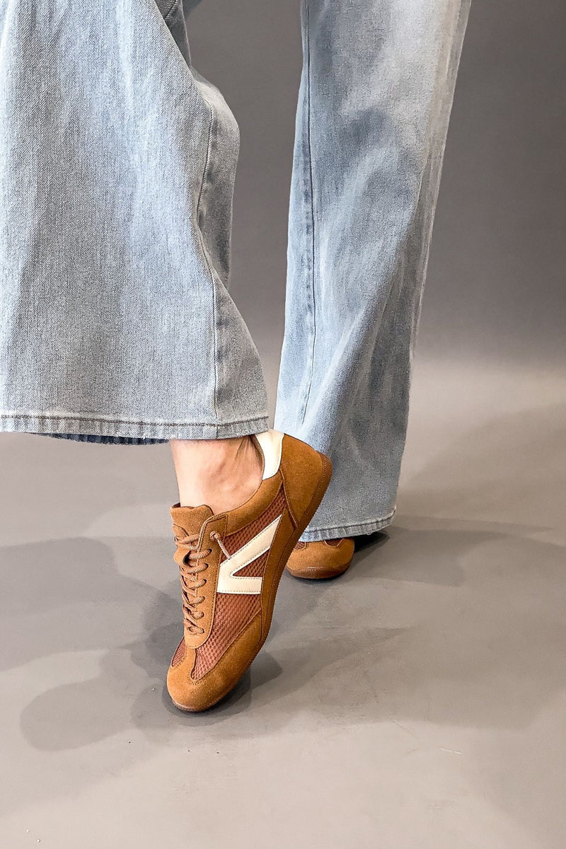 A person wearing light blue wide-leg jeans and camel faux suede sneakers with white side stripes, shown from the calves down against a plain gray studio background.
