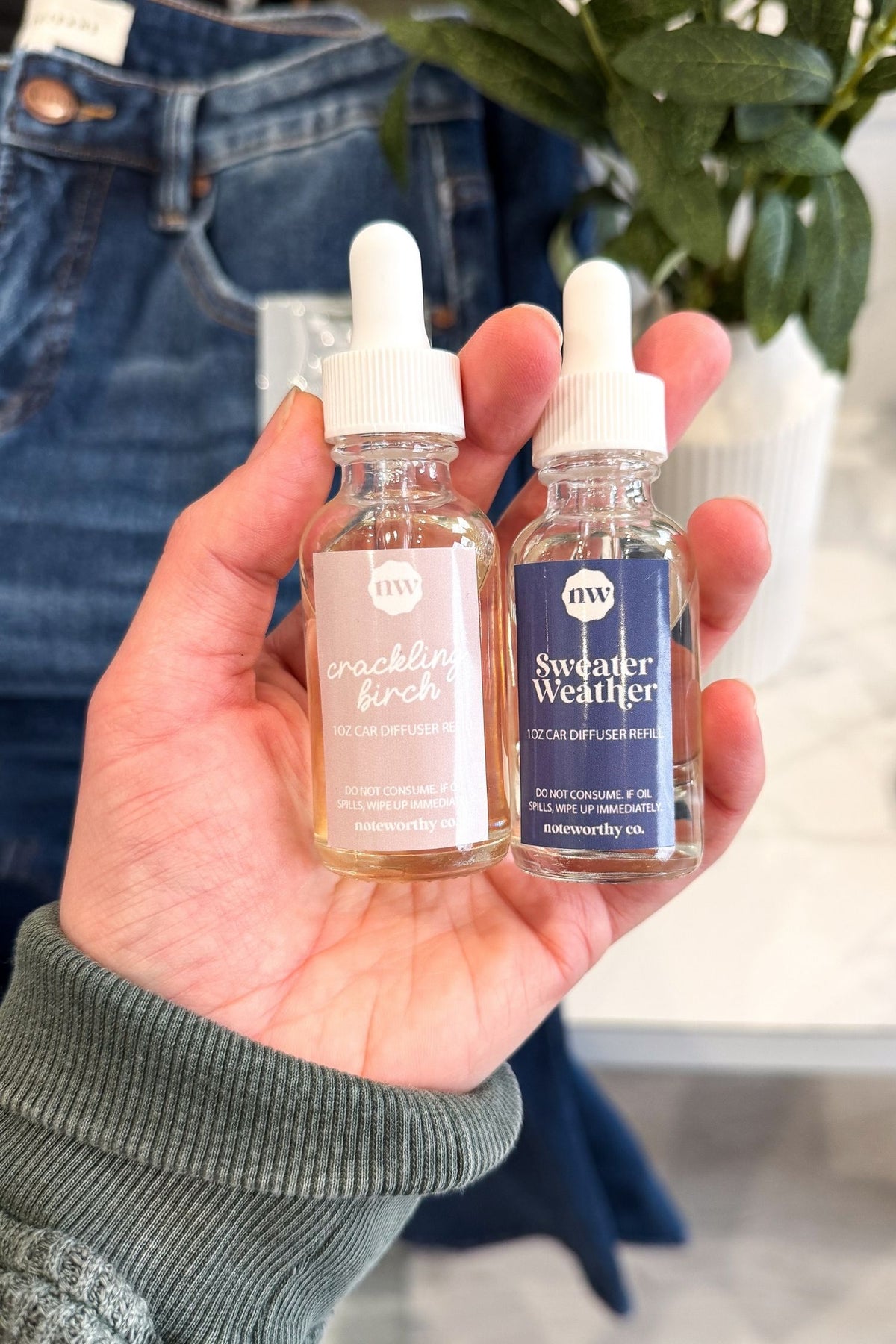 Hand holding two bottles of 'Cracklin' Fir' and 'Sweater Weather' with a blurred background