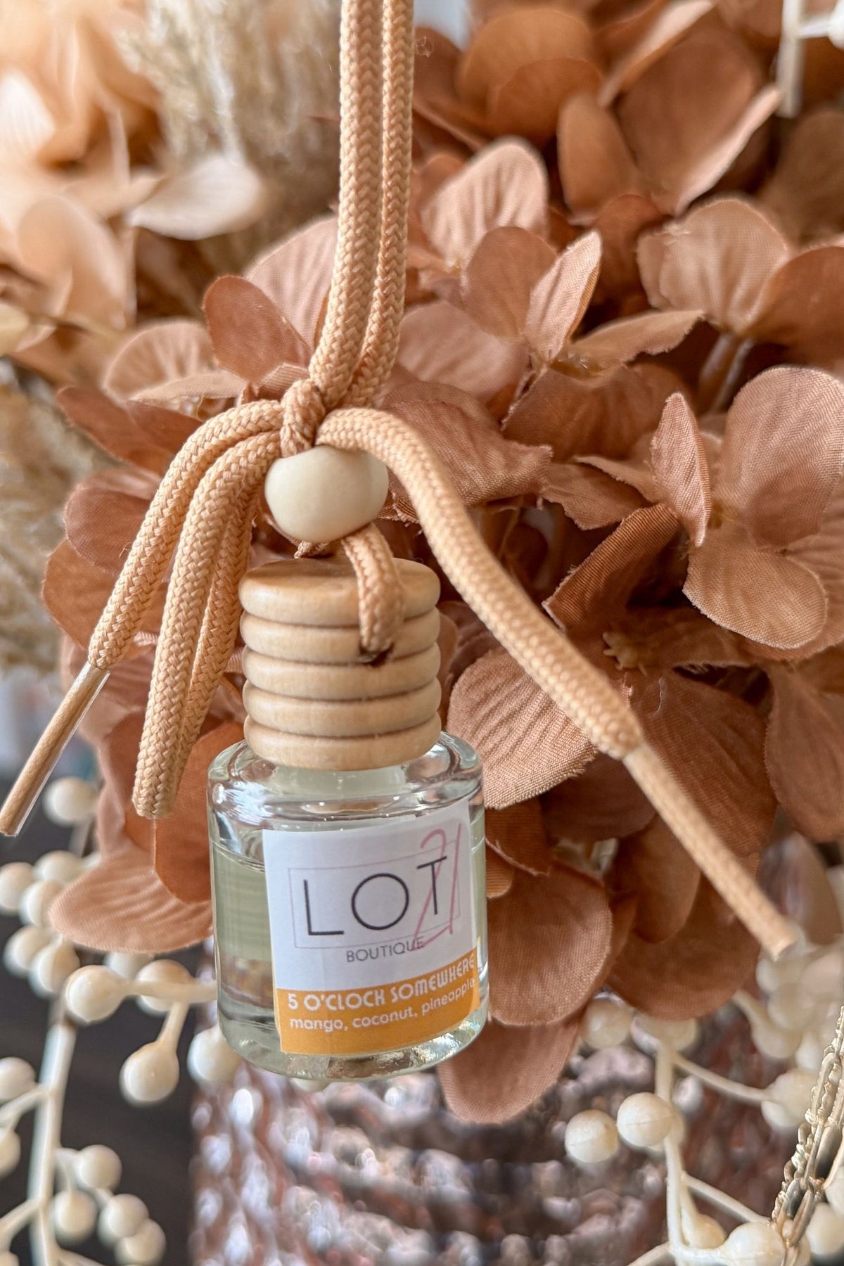 A small clear glass reed diffuser bottle with a light wood cap and a beige hanging cord, shown among dried peach-toned flowers. The bottle label reads LOT21 + 5 O'Clock Somehwere.