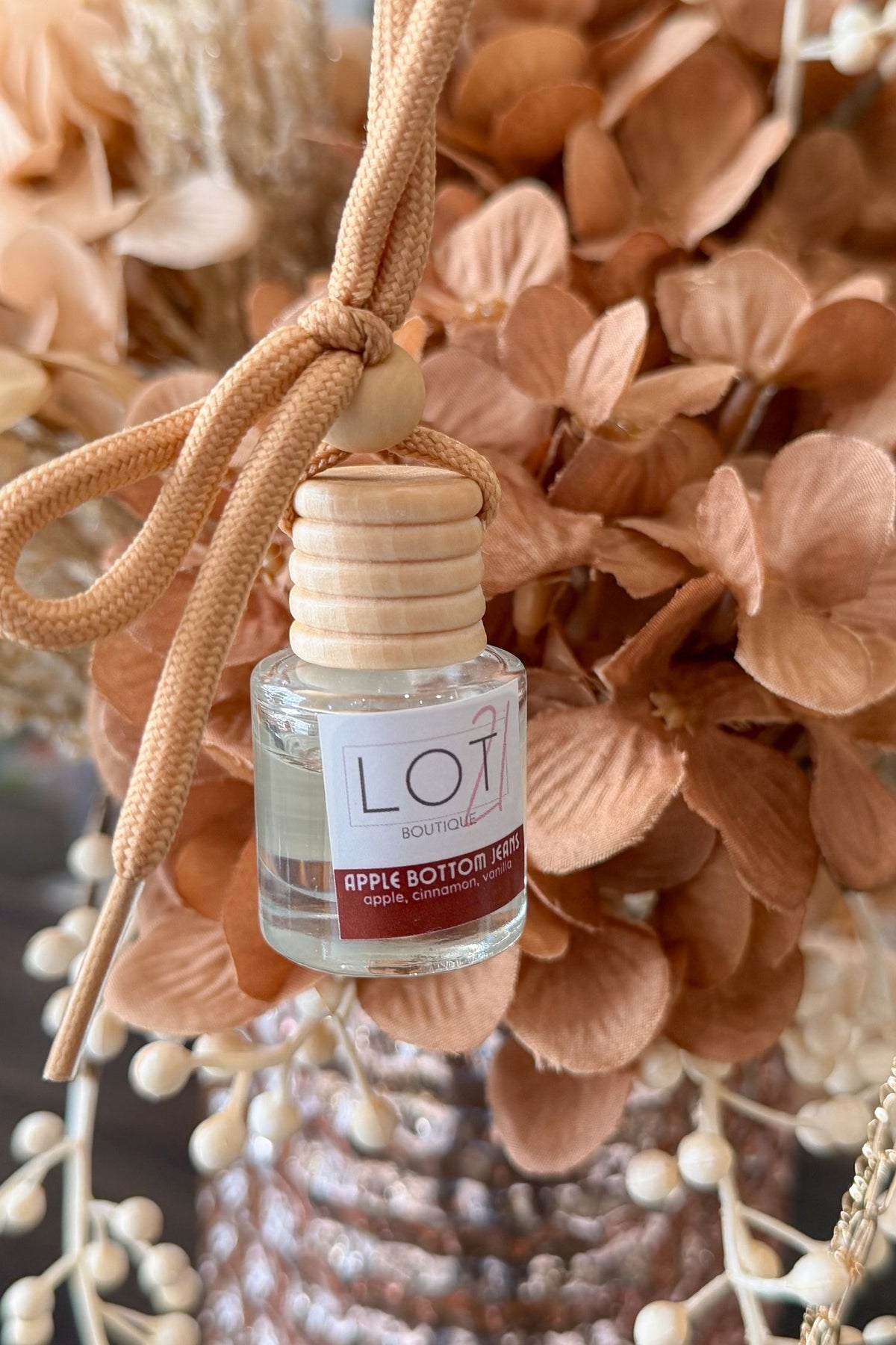A small clear glass reed diffuser bottle with a light wood cap and a tan cord tied around it, shown against a background of dried peach and beige flowers. The label reads LOT21 BOUTIQUE and APPLE BOTTOM JEANS