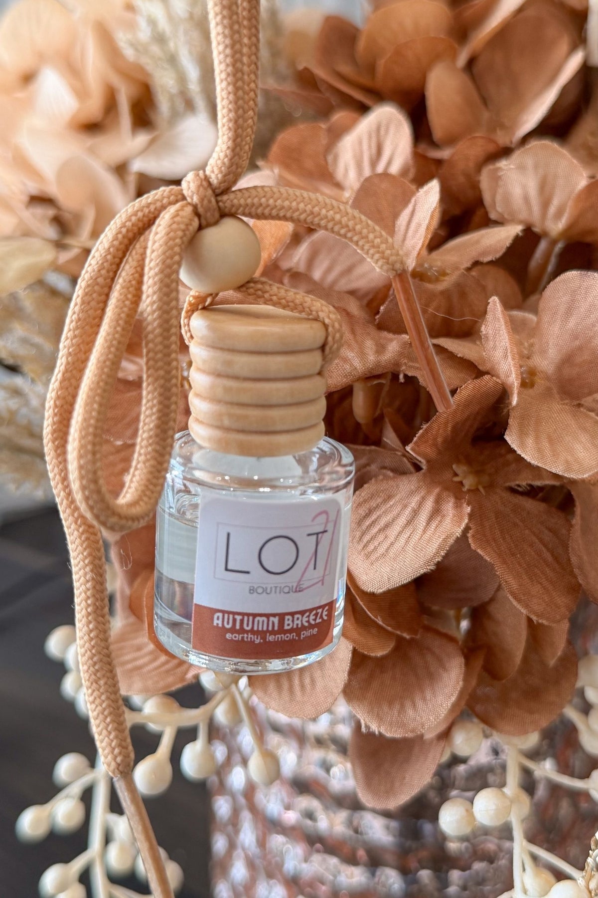A small clear glass reed diffuser bottle with a light wood cap and a beige cord, labeled LOT21 + AUTUMN BREEZE, styled among dried peach and tan flowers.