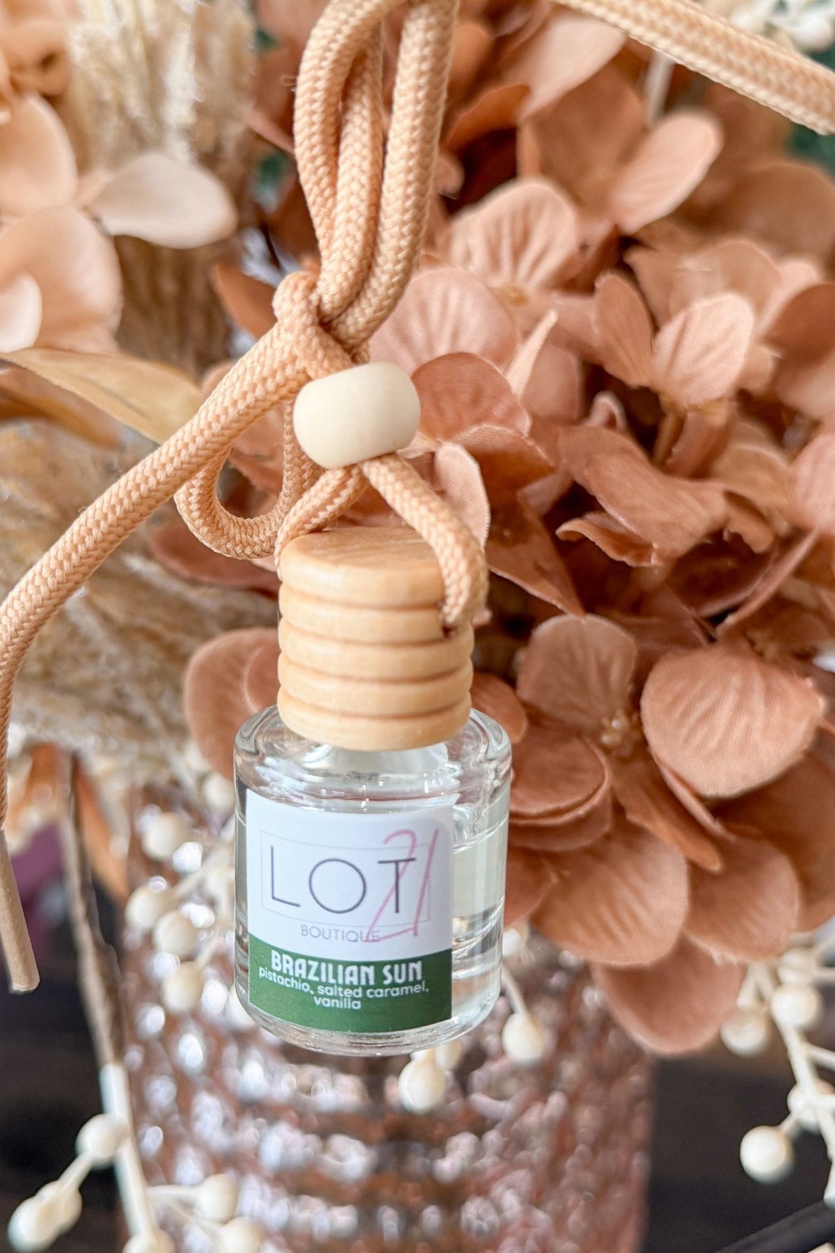 A small clear glass reed diffuser bottle labeled 'LOT21 Brazilian Sun' with a light wood cap and beige hanging cord, shown among dried peach flowers and a textured vase.