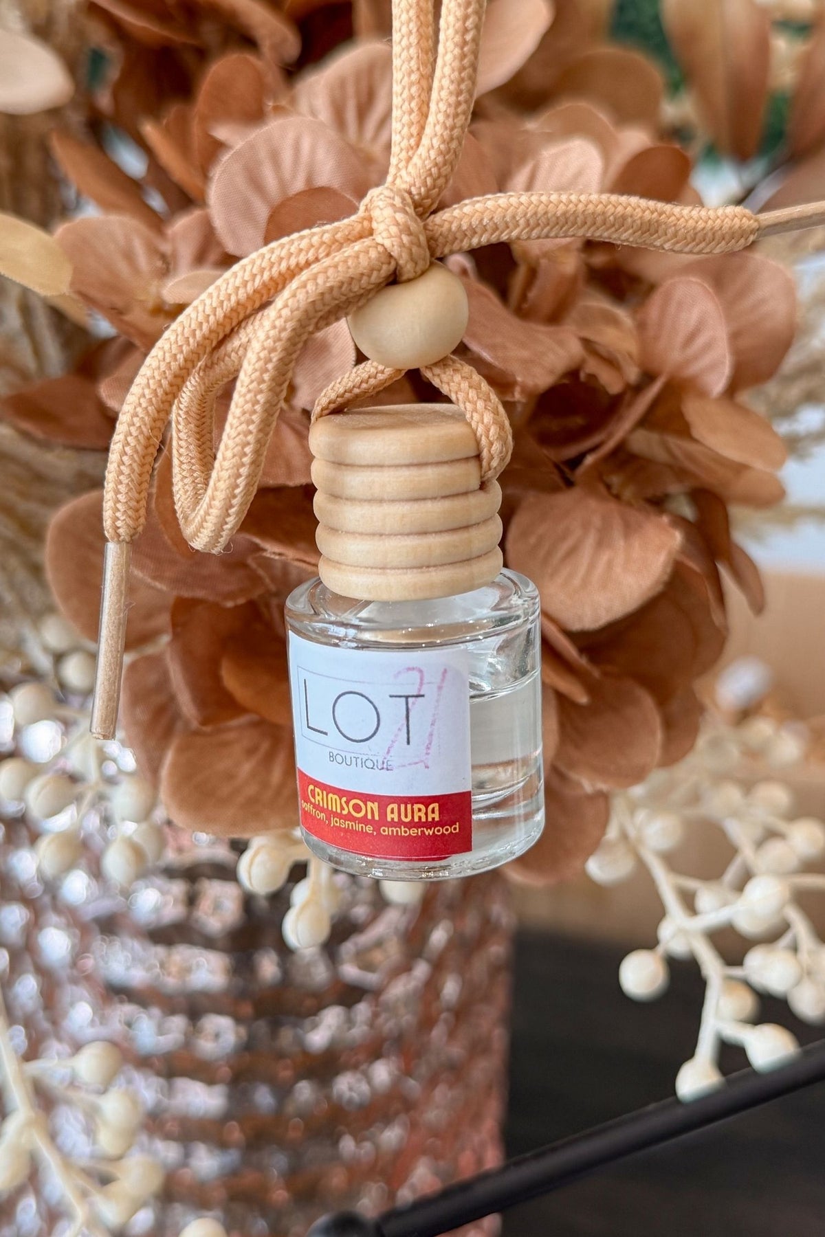 A small clear glass reed diffuser bottle with a light wood cap and a beige braided hanging cord, shown in front of dried tan flowers. The label reads LOT21 Cinnamon Aura.