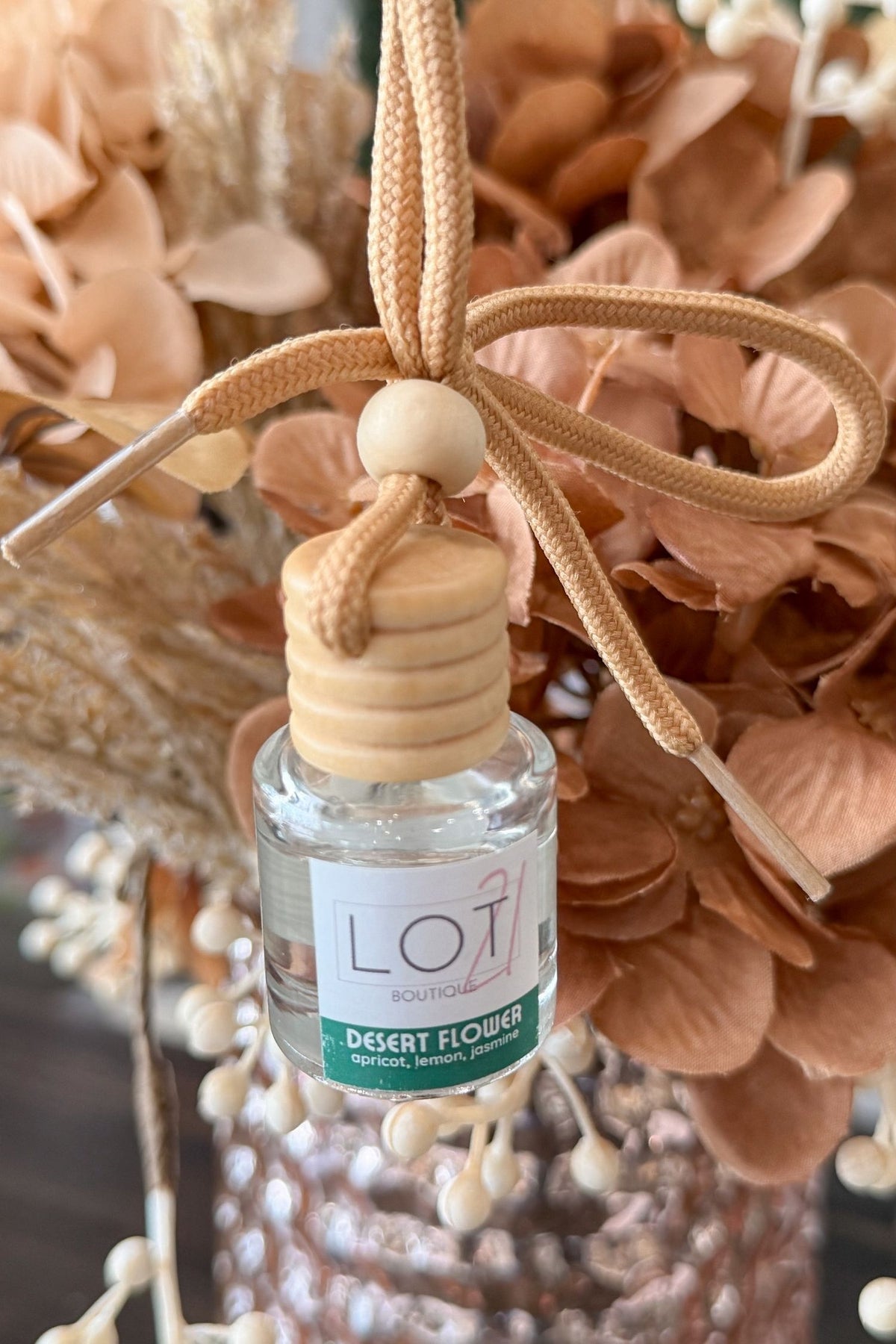 A small clear glass reed diffuser bottle with a beige wooden cap and a beige hanging cord tied in a bow, placed in front of dried peach-toned flowers. The label reads LOT21 and Desert Flower.
