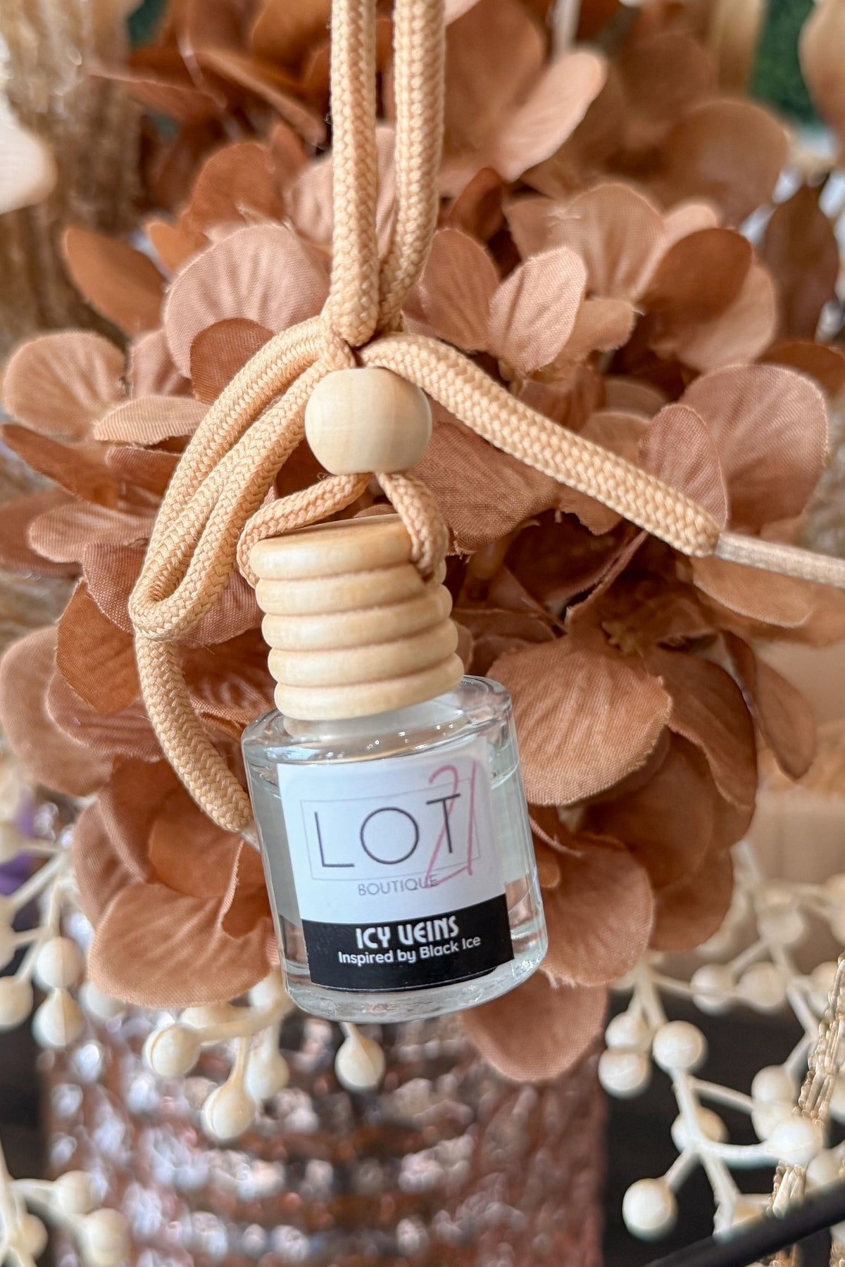 A reed diffuser bottle with a beige woven hanging cord, wooden diffuser top, and a clear glass bottle labeled 'LOT21 + Icy Veins', set against dried peach-colored flowers.