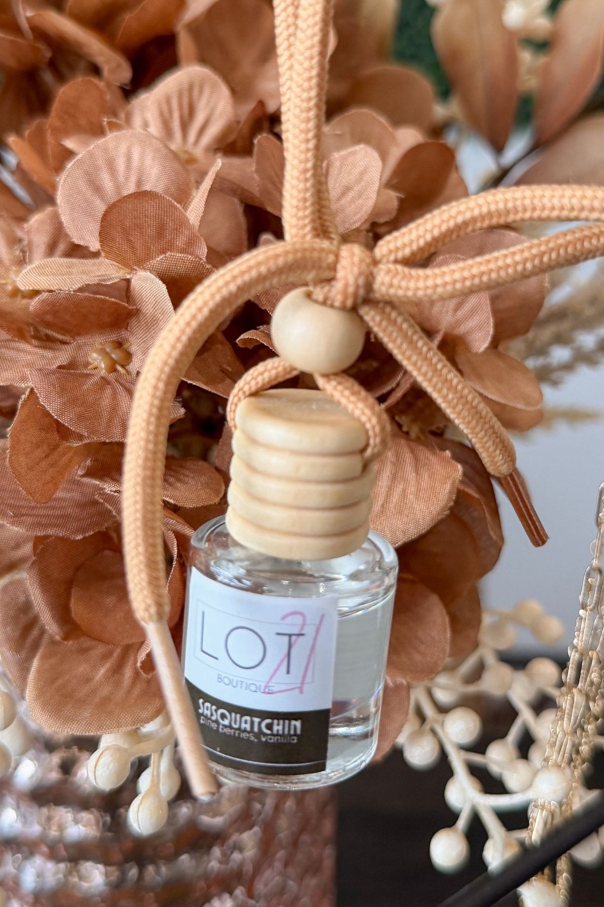 A clear glass reed diffuser bottle with a light wood cap and a beige braided hanging cord, shown in front of dried peach-toned flowers. The bottle label reads LOT 21 and SASQUATCHIN