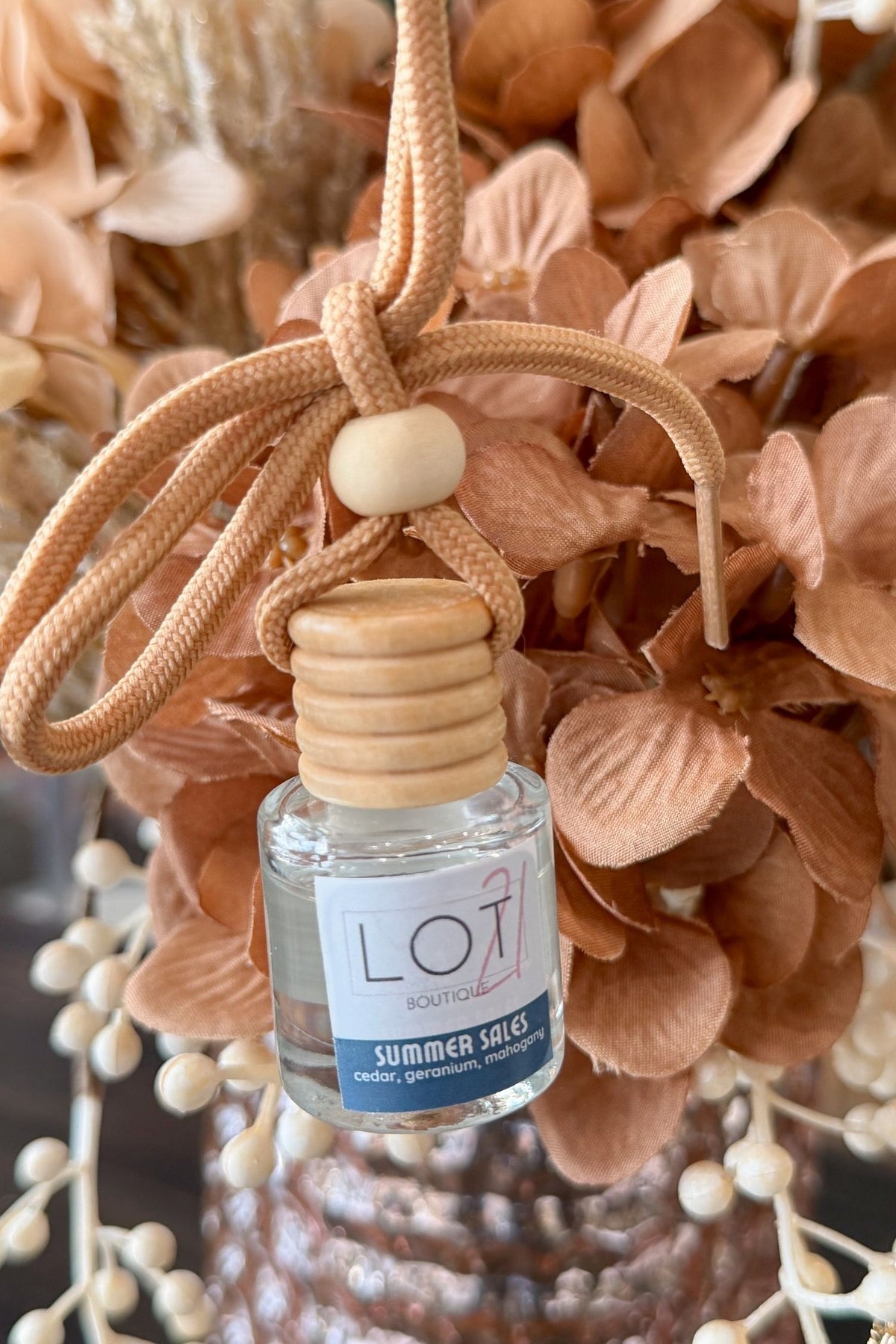 A small clear glass reed diffuser bottle labeled 'LOT scents summer sales' hanging from a tan braided cord with a wooden stopper and decorative beads, set against dried peach-toned flowers.