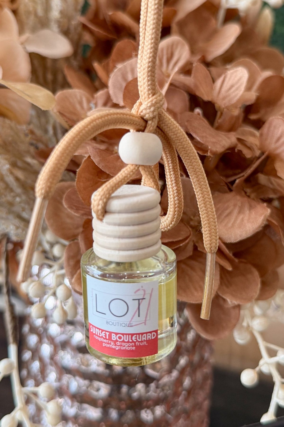 A small clear glass reed diffuser bottle with pale yellow liquid, a white label reading LOT21 + Sunset Boulevard, and a natural wood diffuser top, hanging from a tan braided cord with a knot and white bead in front of dried brown flowers.