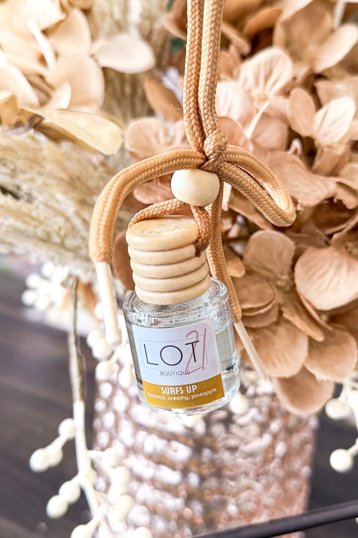 A close-up of a reed diffuser bottle labeled 'LOT 21 Surfs Up' with a beige cord and wooden cap, surrounded by dried beige flowers and a textured vase.