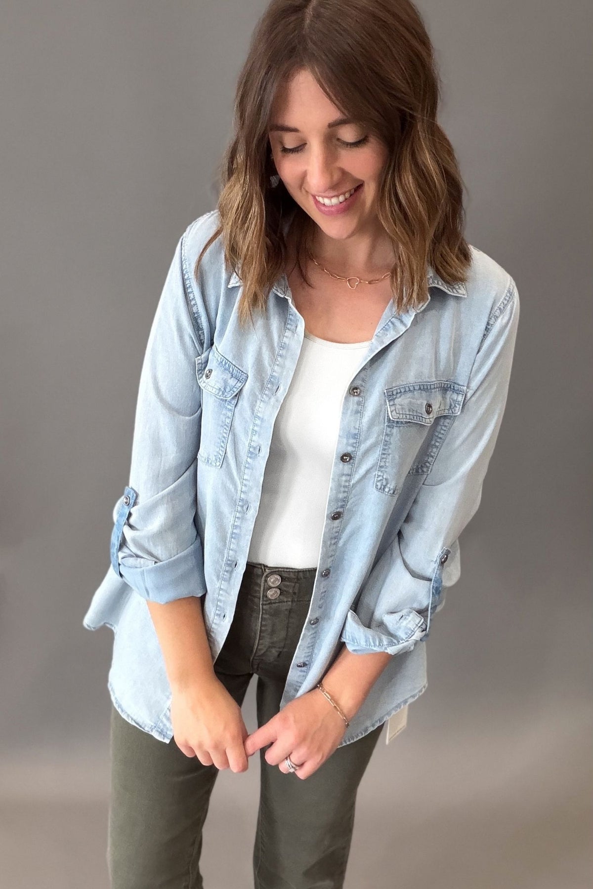 a woman that has shoulder length hair wearing a chambray button front button up with the sleeves rolled up, paired with a white tank and olive mica denim