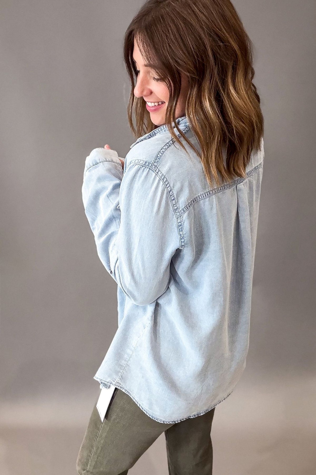 A woman wearing a light-wash blue denim shirt and olive green pants, shown from a side-back angle against a neutral gray studio background.
