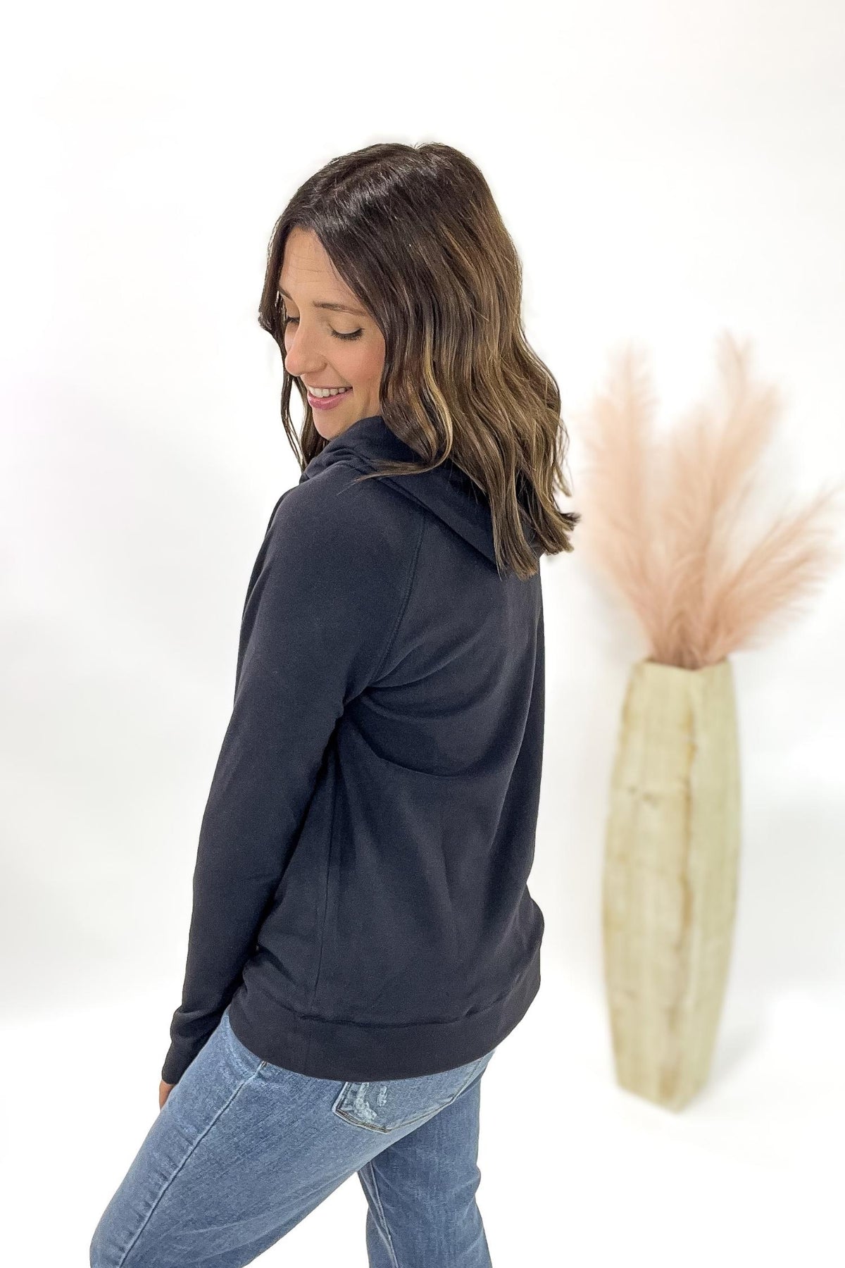 Woman wearing a charcoal hoodie and jeans standing next to a vase with dried plants on a white background