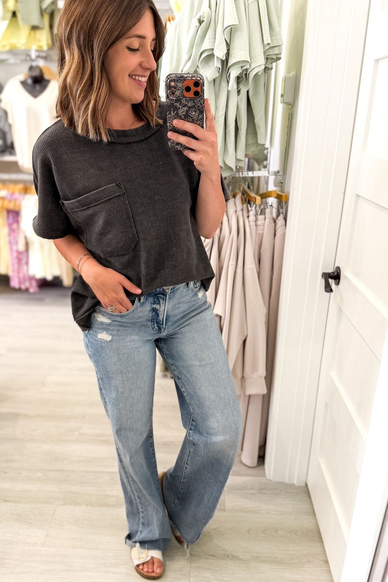 A woman takes a mirror selfie in a clothing store, wearing a dark charcoal gray short-sleeve t-shirt with a chest pocket and light-wash blue jeans.