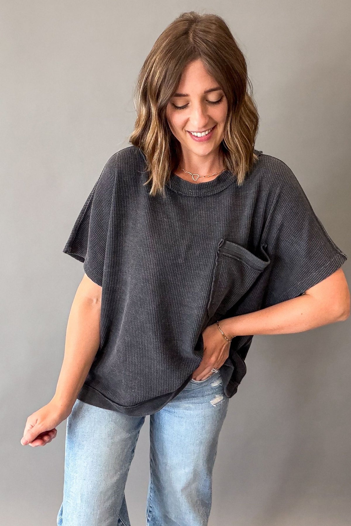 A woman wearing a dark charcoal gray short-sleeve waffle-knit top with a chest pocket, paired with light blue jeans, standing against a plain gray background.