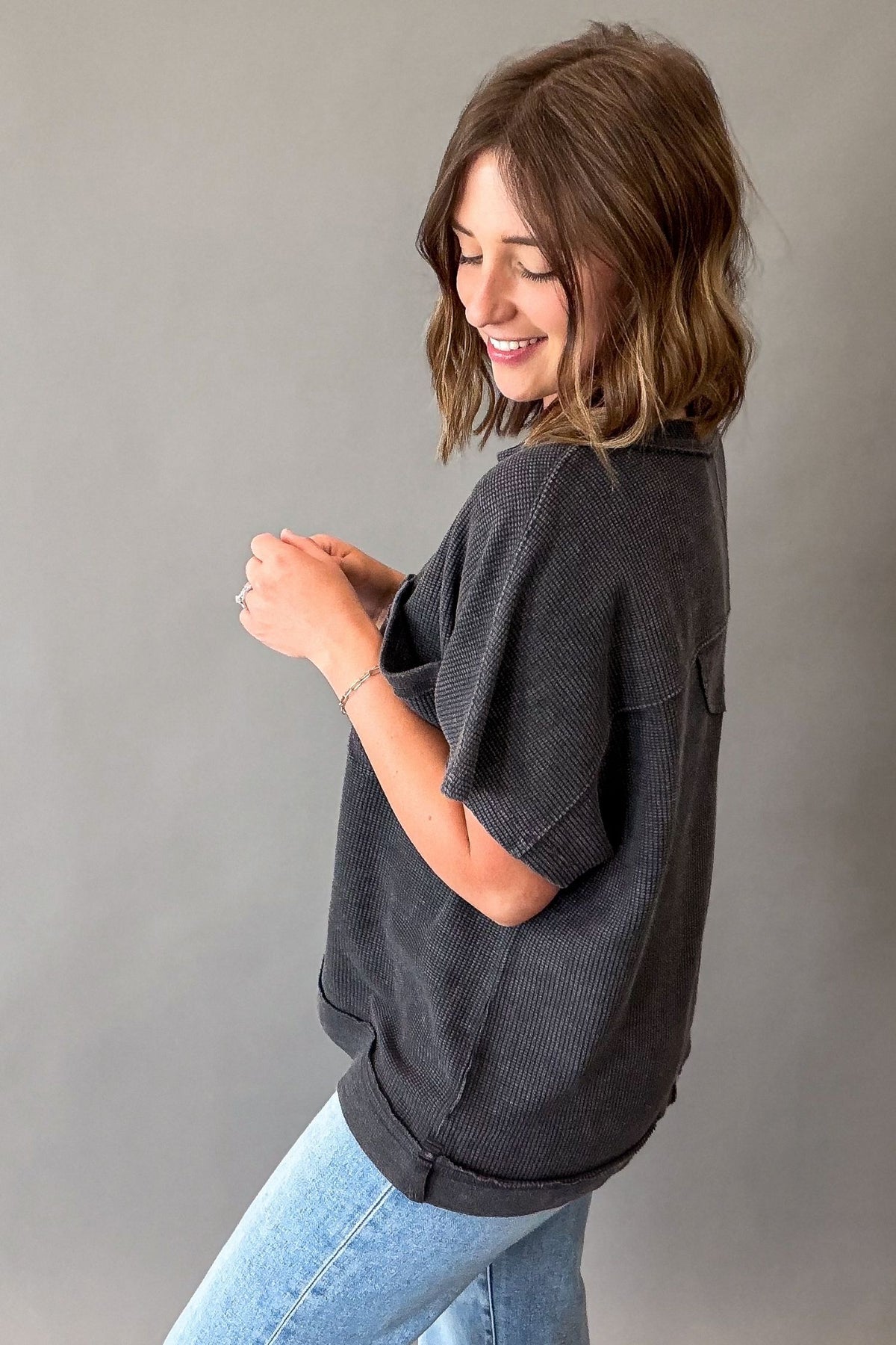 A woman wearing a dark charcoal gray ribbed short-sleeve top and light blue jeans, posing against a solid gray studio background.
