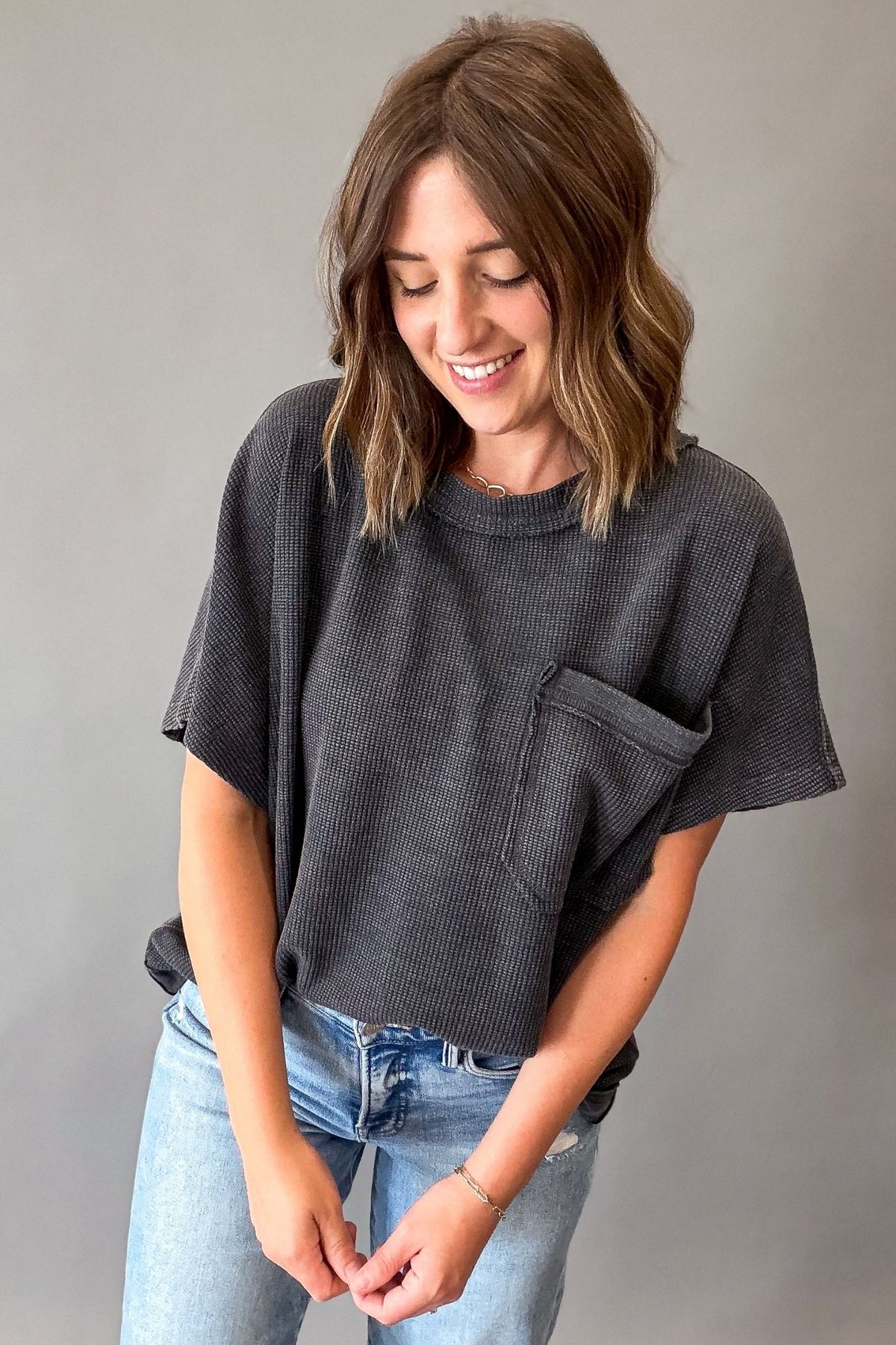 A woman wearing a dark gray ribbed short-sleeve t-shirt with a chest pocket and light blue jeans, standing against a solid gray background.