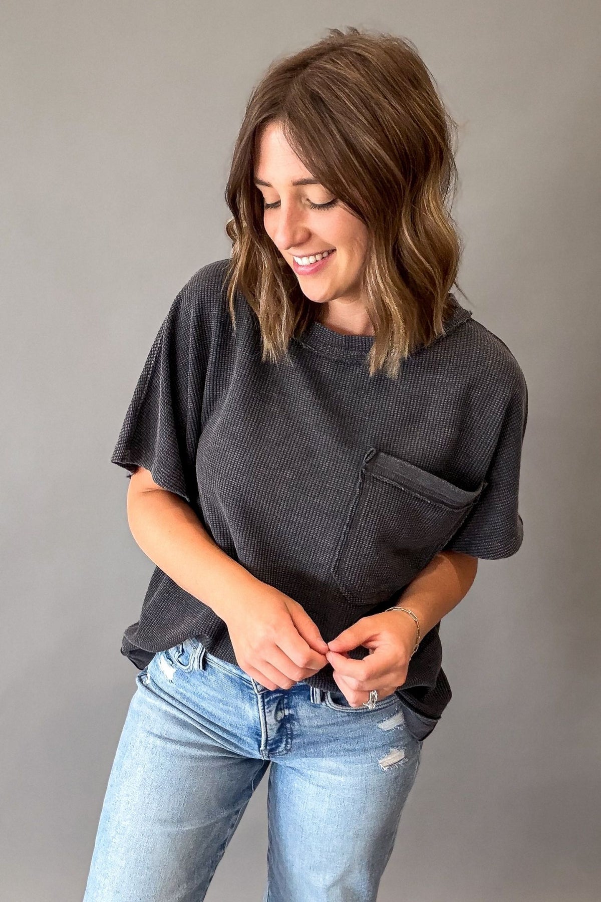 A woman with shoulder-length brown hair wearing a dark charcoal gray ribbed short-sleeve t-shirt with a chest pocket and light-wash blue jeans, standing against a solid gray background.