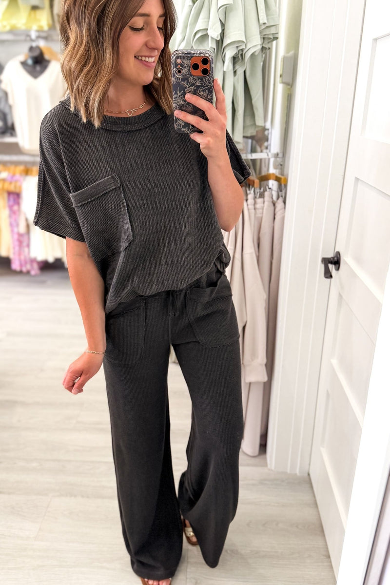 A woman takes a mirror selfie in a clothing boutique wearing a dark charcoal gray short-sleeve t-shirt with a chest pocket and matching dark gray wide-leg pants.