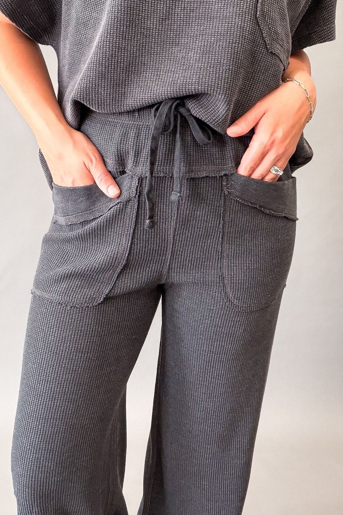 A close-up of a person wearing dark gray ribbed lounge pants with a drawstring waist and large front patch pockets, paired with a matching gray top.