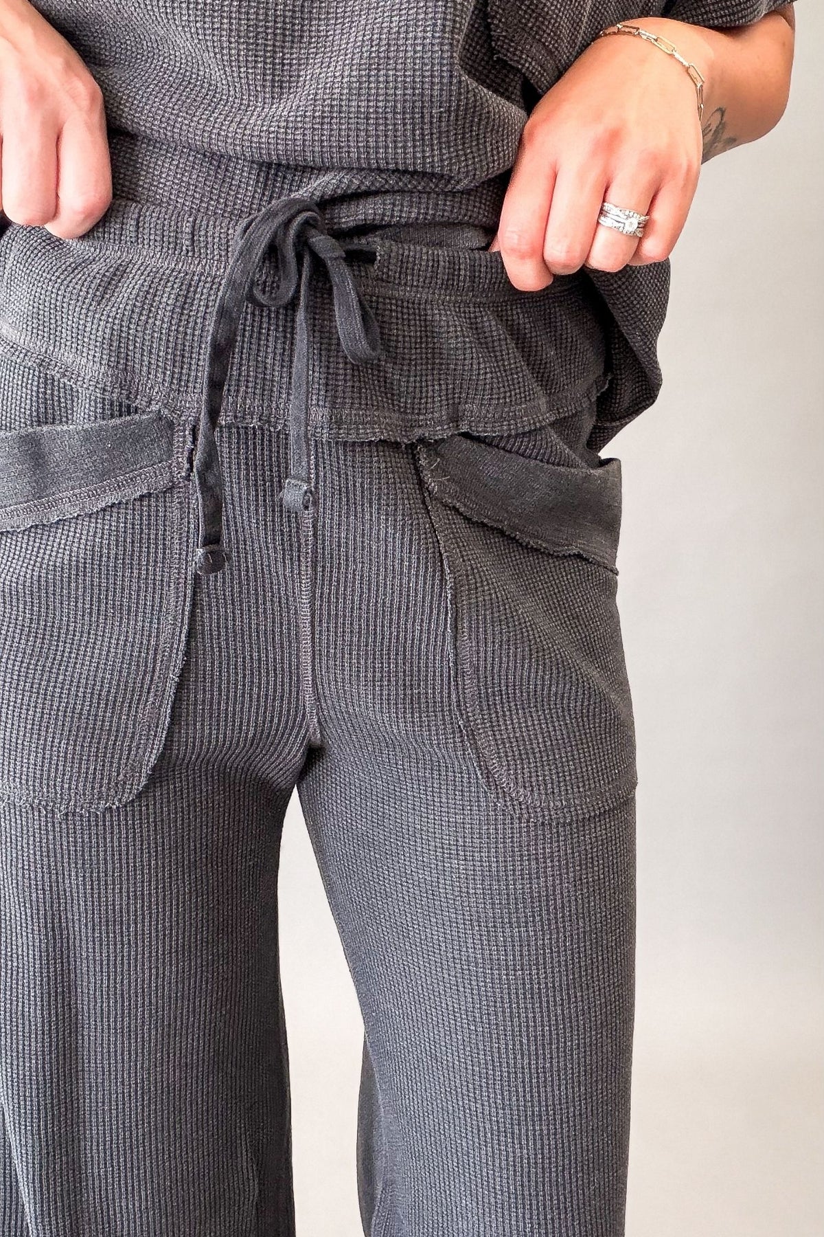 Close-up of a person wearing dark gray ribbed lounge pants with a drawstring waist and large front patch pockets, paired with a matching top.