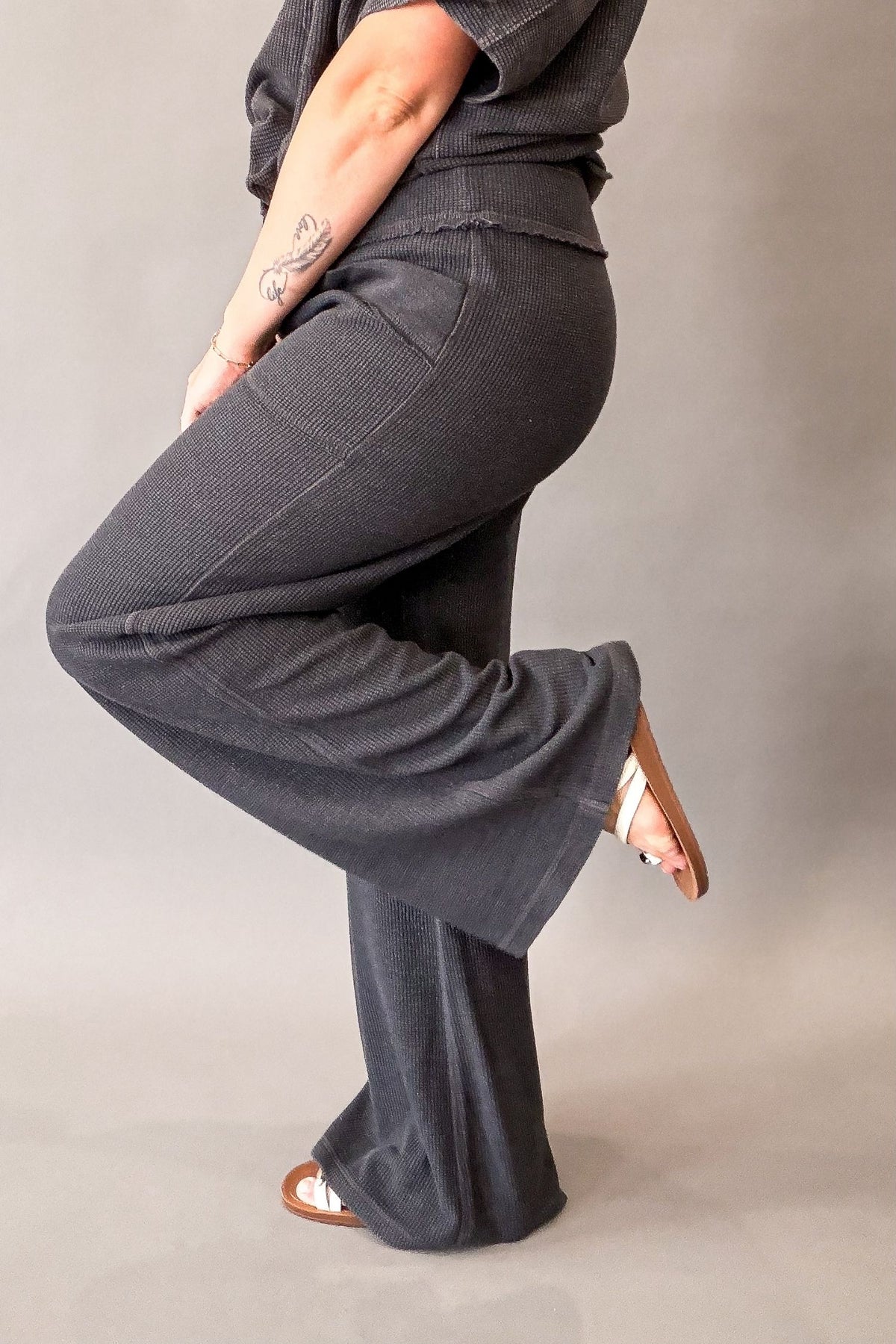 A person wearing dark charcoal gray wide-leg pants and a matching dark top, shown from the waist down in a studio setting against a plain gray backdrop.