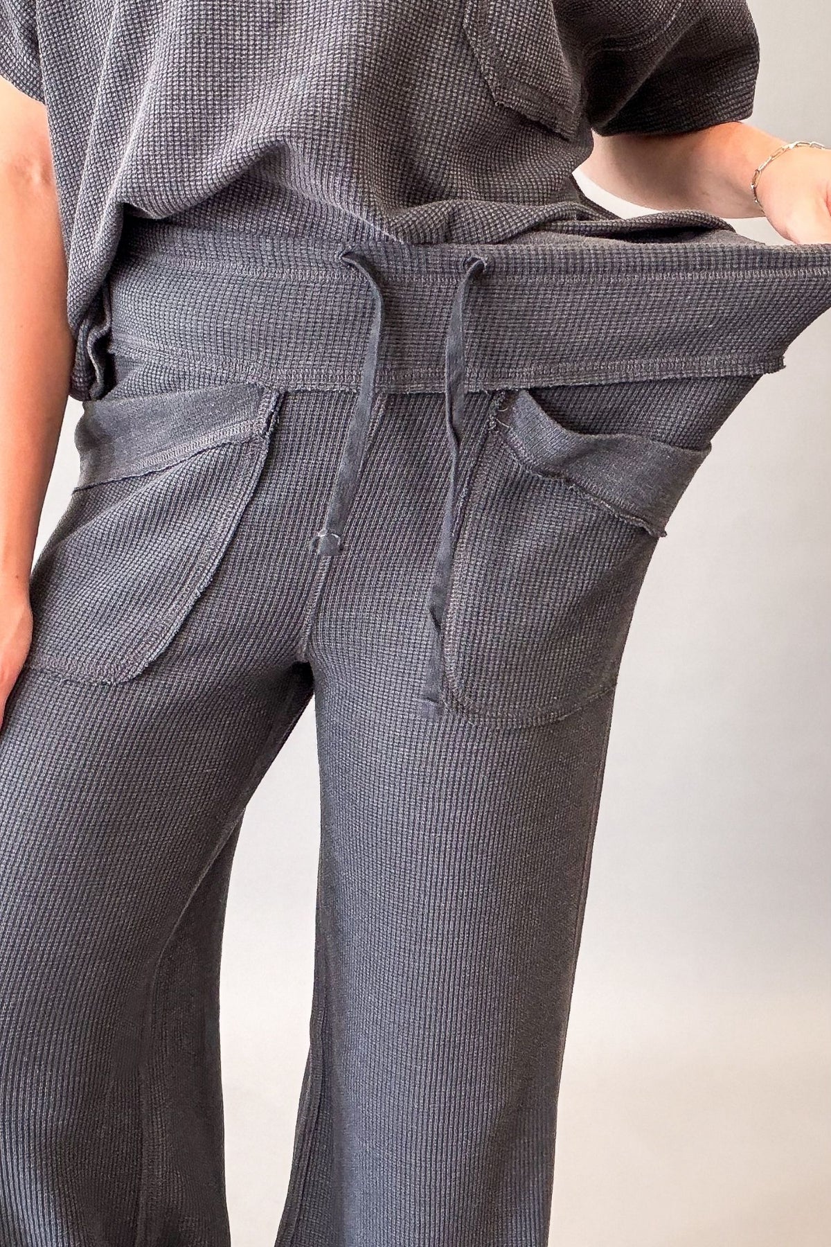 Close-up of a person wearing dark gray ribbed wide-leg pants with a drawstring waistband and large front patch pockets, holding the waistband open to show the fabric stretch.