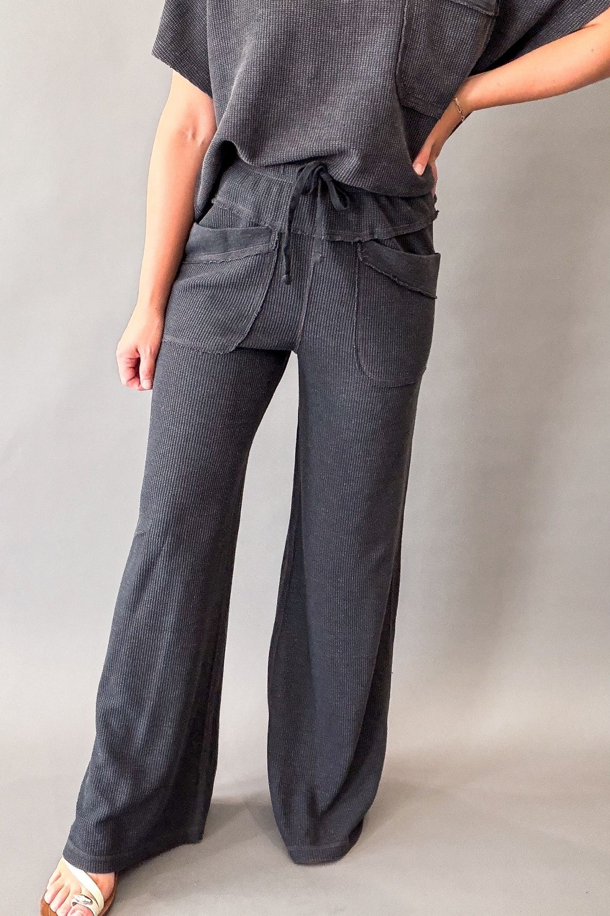 A person wearing dark charcoal gray wide-leg pants with a drawstring waist and large front patch pockets, paired with a matching ribbed top, photographed against a plain light gray studio background.