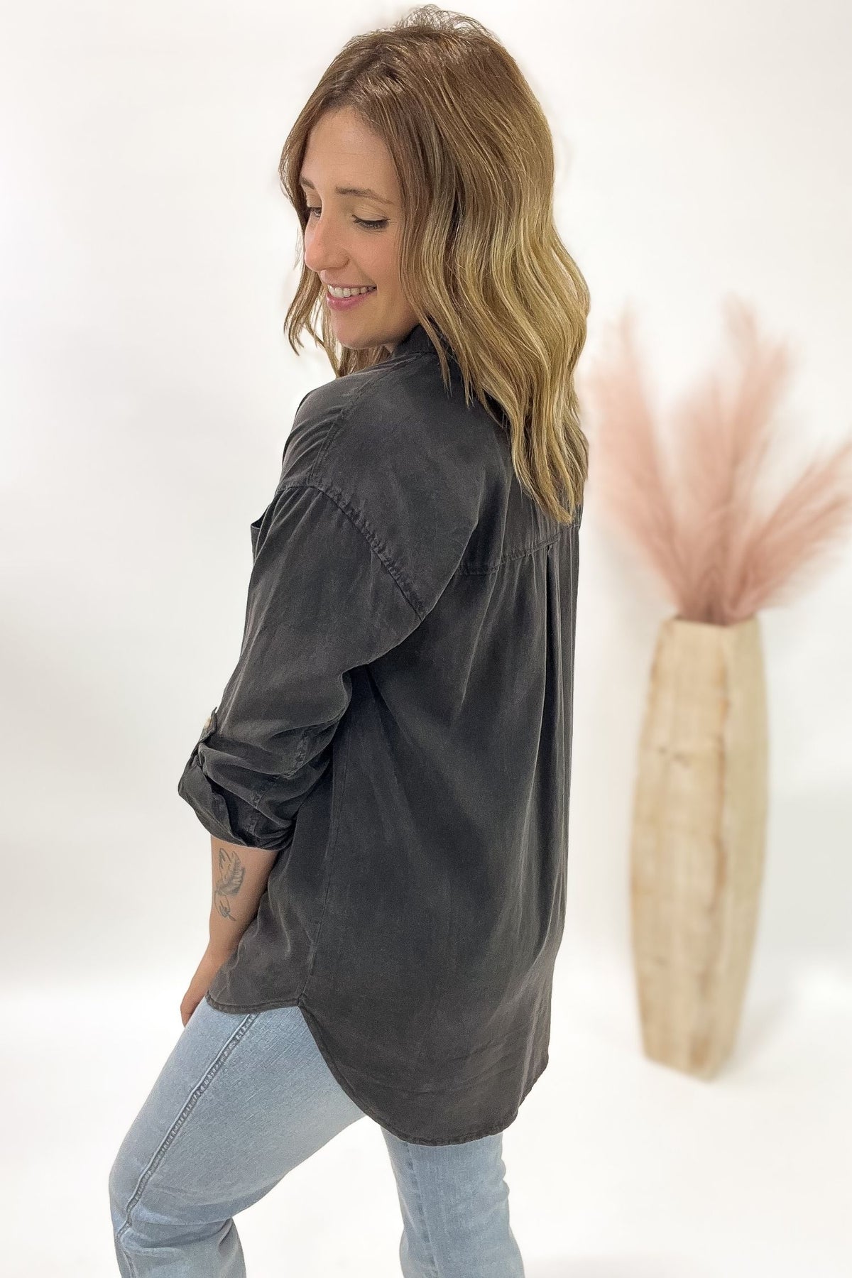Woman wearing a dark gray oversized shirt and light blue jeans standing against a white background with a vase of dried pink flowers.