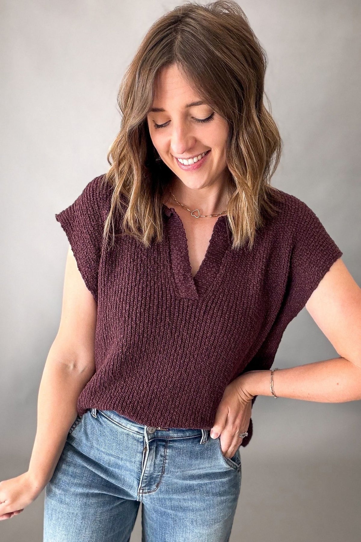 A woman wearing a short-sleeve ribbed knit top in a deep plum purple color, paired with blue jeans, posing against a neutral gray background.
